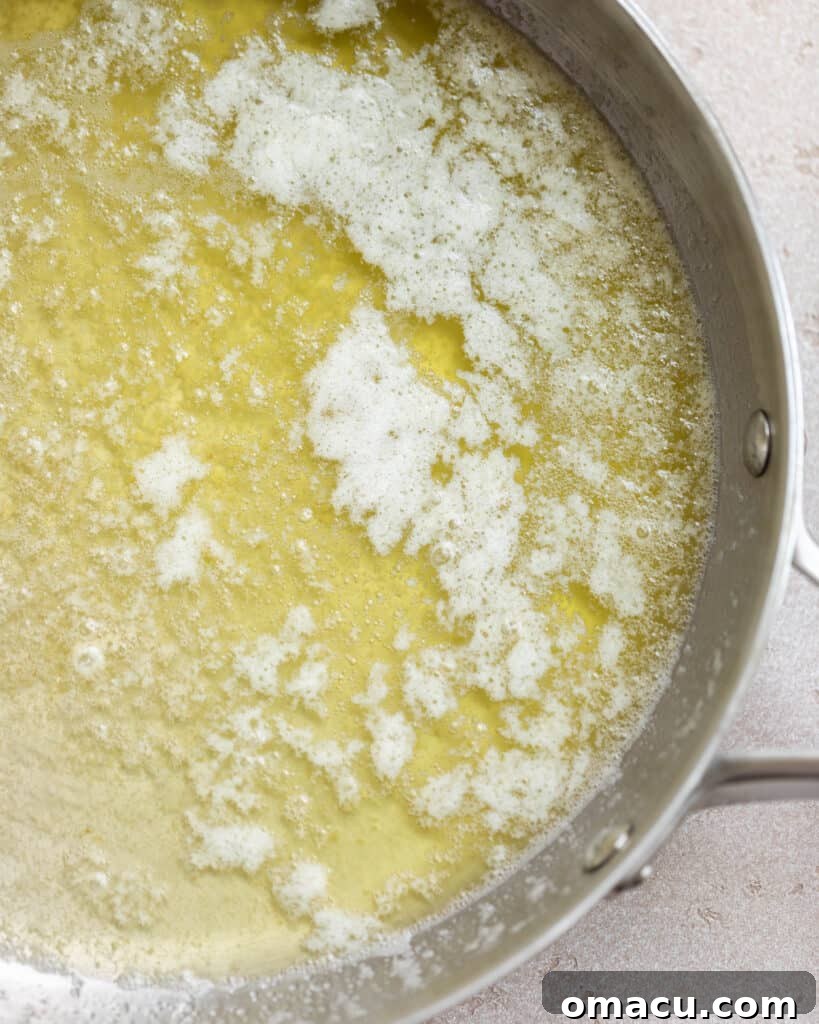 Melted butter in a pan, actively foaming and beginning to show tiny brown specks at the bottom, indicating the start of the browning process.