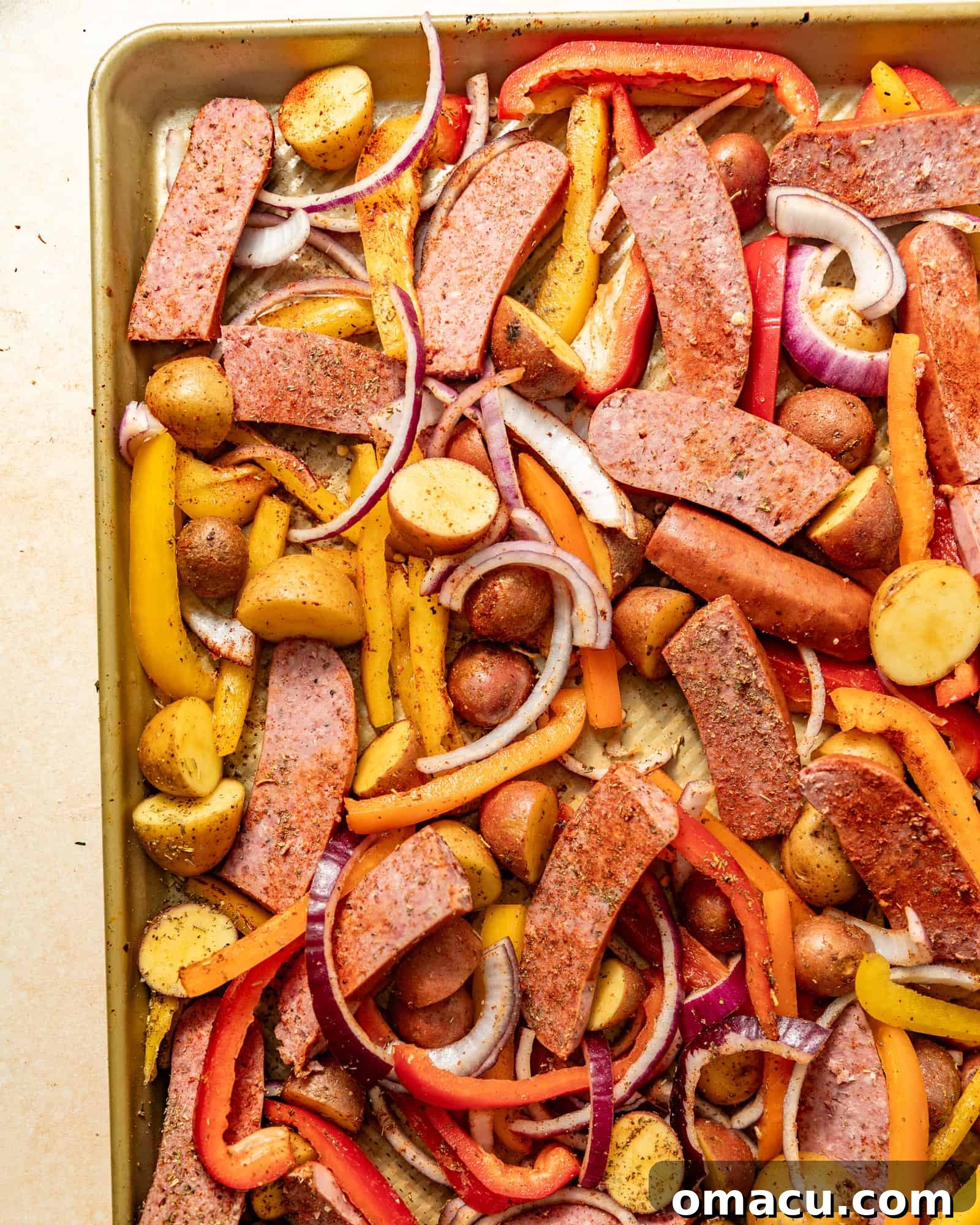 Sausages, peppers, onions, and potatoes seasoned with Italian seasoning and smoked paprika, drizzled with olive oil on a sheet pan, ready for the oven.