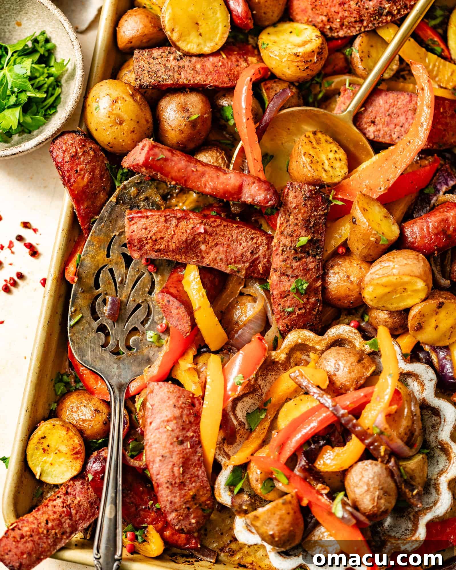 A sheet pan filled with roasted sausages, peppers, onions, and potatoes, alongside a small bowl of fresh parsley for garnish.