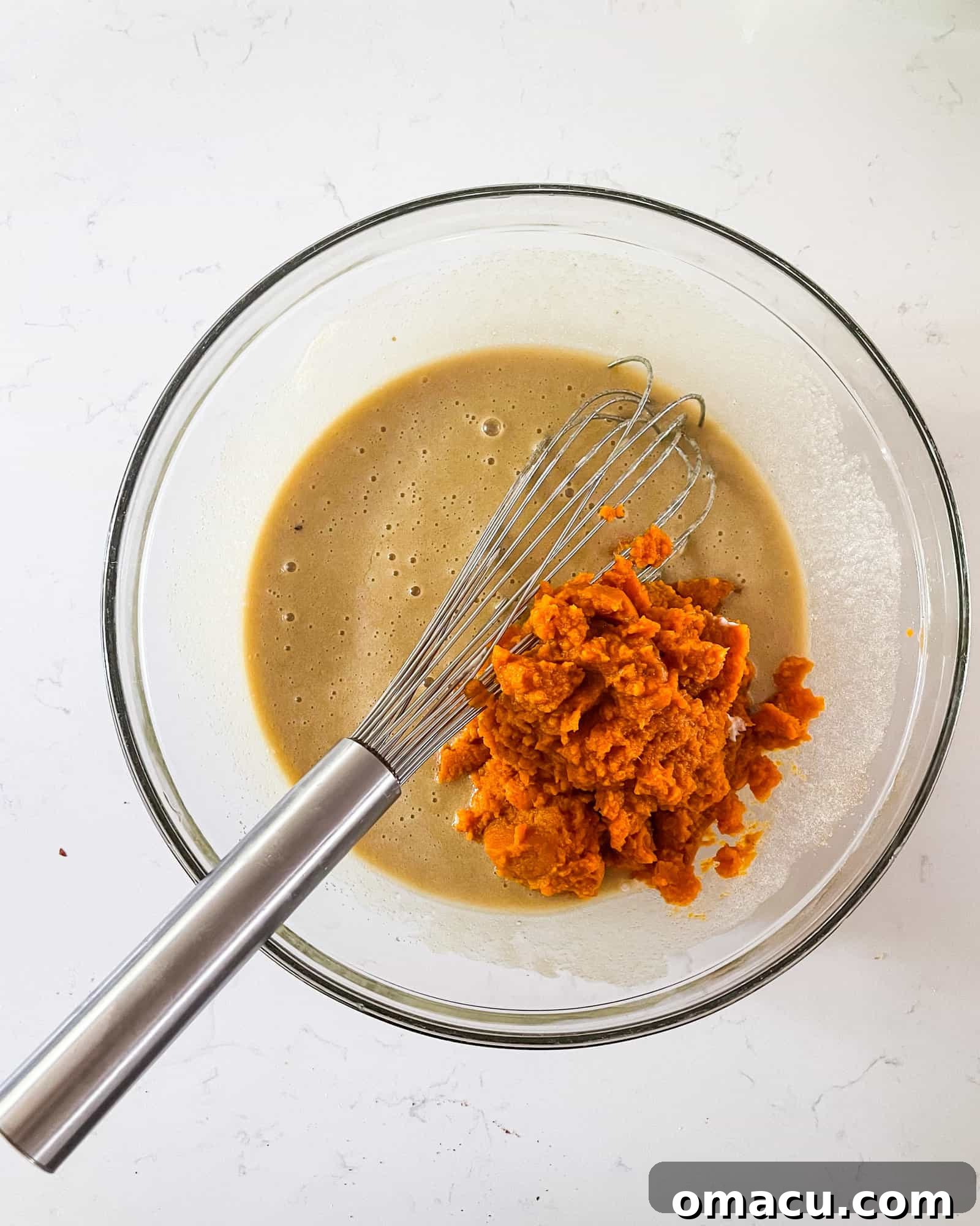 Pureed pumpkin being added to the mixed wet ingredients in a bowl, creating a rich, autumnal batter with a whisk nearby.
