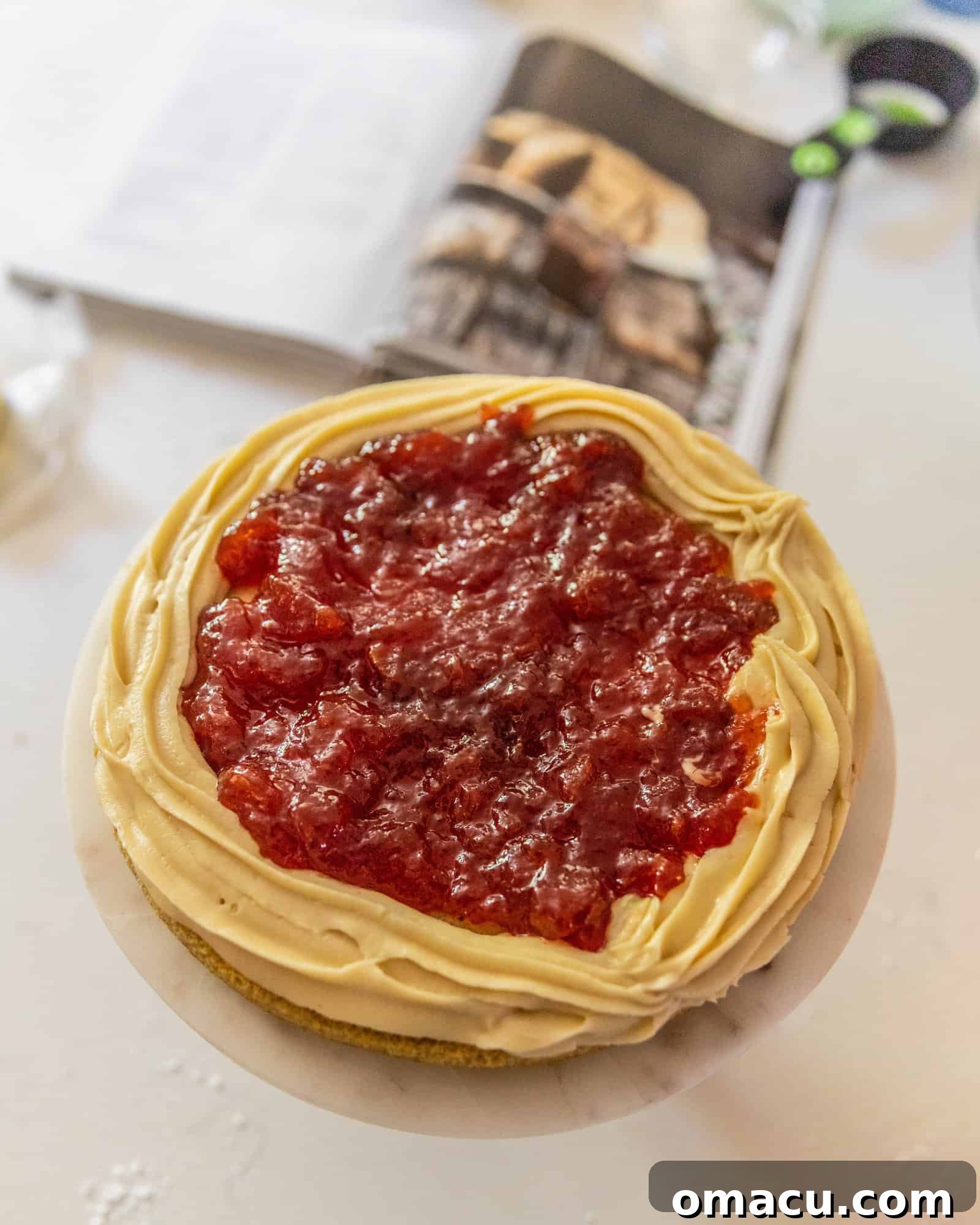 A two-layer cake being assembled, showing the piped buttercream dam and strawberry jam filling before the top layer is added.