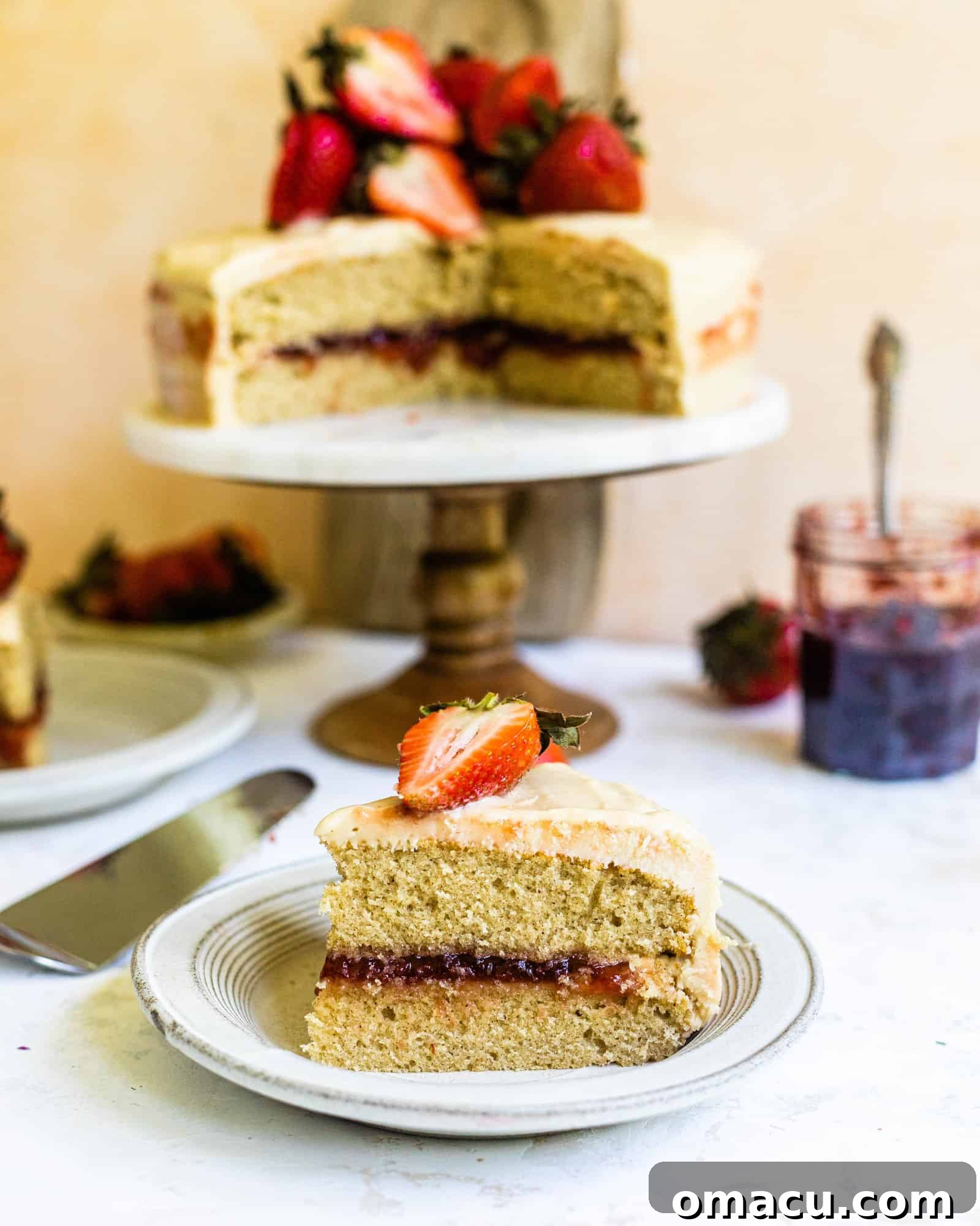 A slice of Strawberry Jam Cake on a plate, with the larger cake beautifully presented on a stand in the background, inviting indulgence.