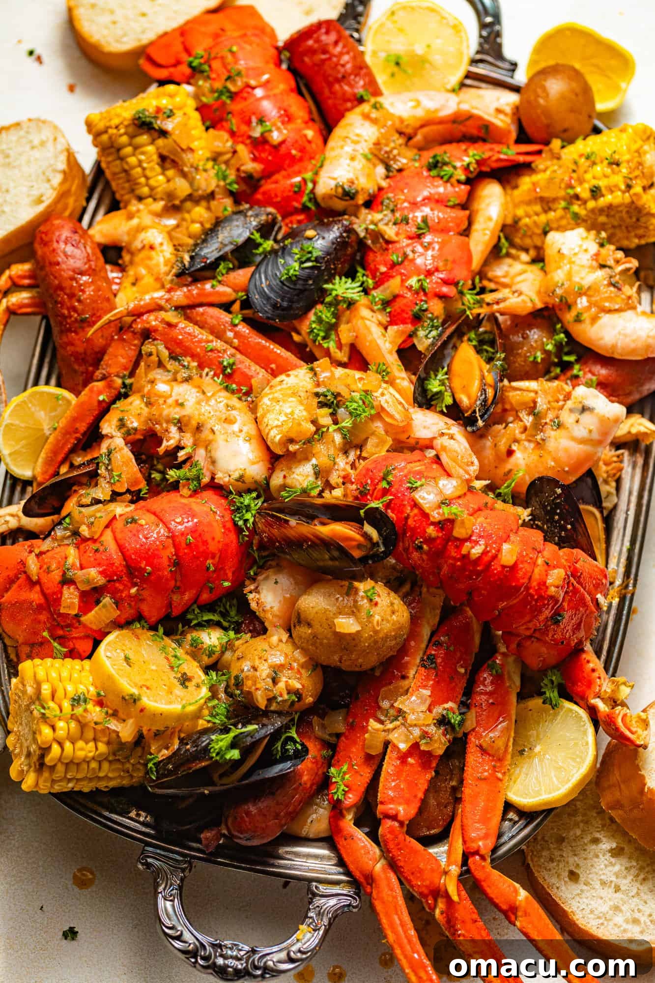 A lavish spread of crab legs, lobster tails, corn cobs, sliced sausage, tender potatoes, fresh clams, plump mussels, and jumbo shrimp, all presented on a gleaming silver serving tray, drenched in rich seafood boil sauce and garnished with vibrant fresh parsley.