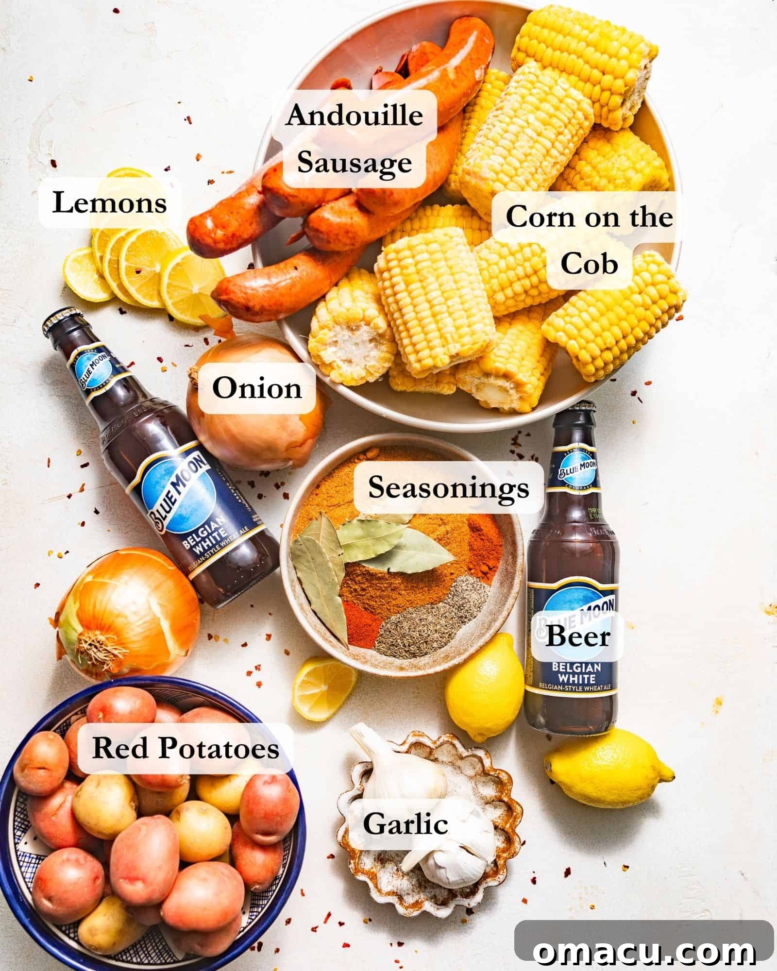 A visual display of essential ingredients for a Cajun seafood boil: beer, Old Bay seasoning, fresh corn, whole onions, smoked sausage, small red potatoes, bright lemons, garlic cloves, bay leaves, and a blend of Cajun seasoning, all arranged on a pristine white surface.