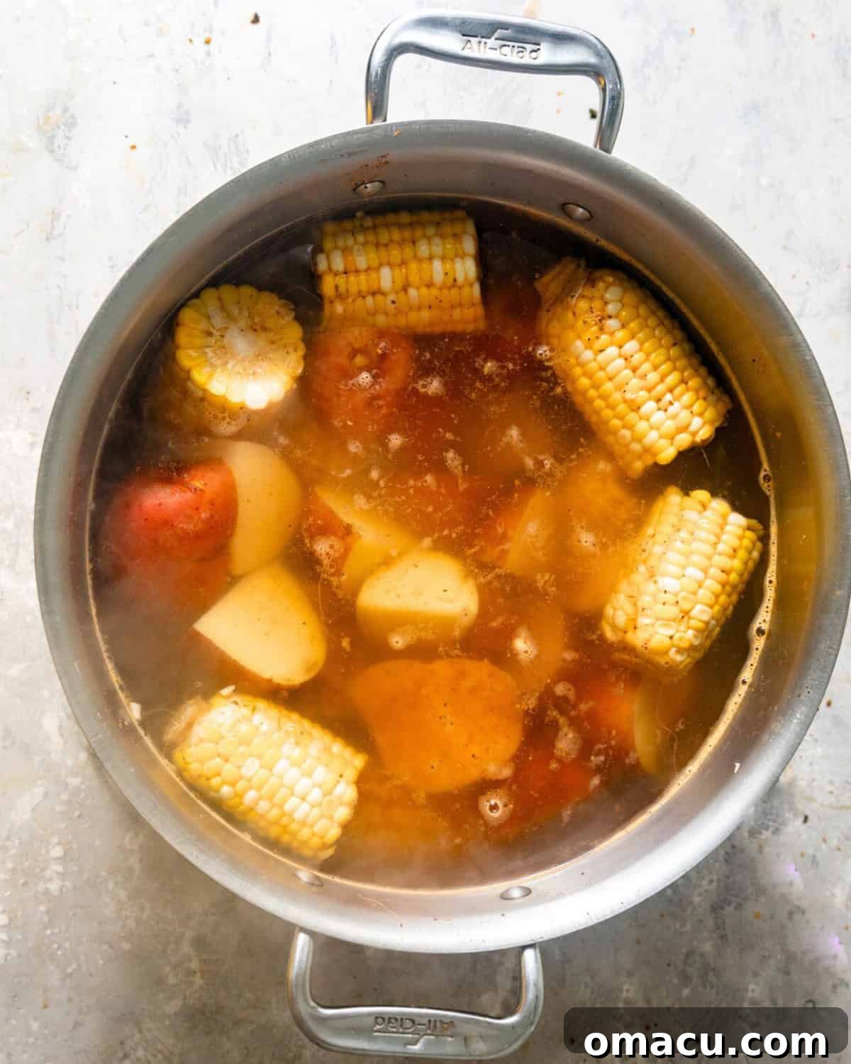 Overhead view of corn, sausages, and halved red potatoes simmering in a large pot of seasoned broth, preparing for the seafood boil.