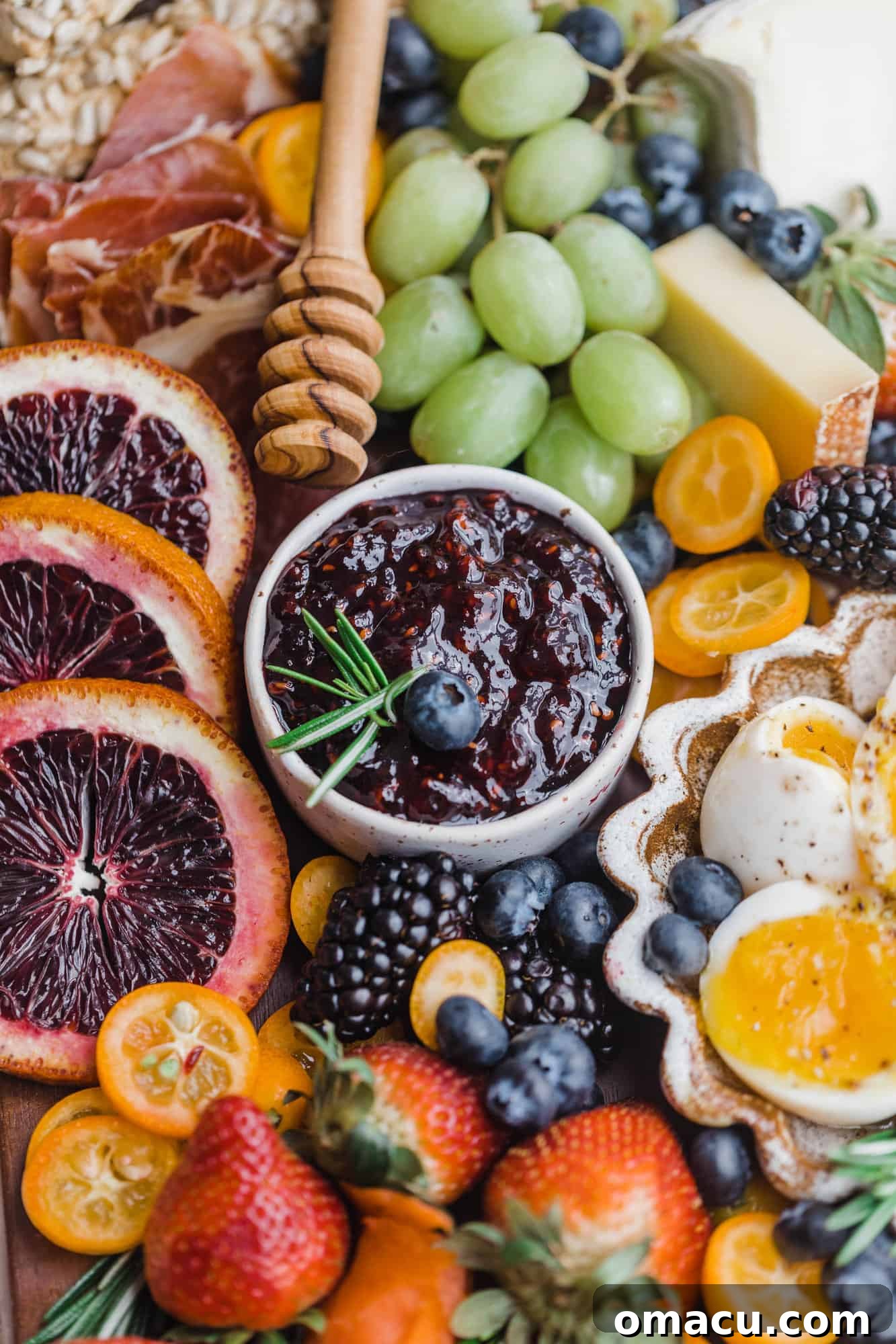 Raspberry preserves in a small bowl, artfully surrounded by colorful sliced blood oranges, fresh strawberries, and green grapes on a wooden board.