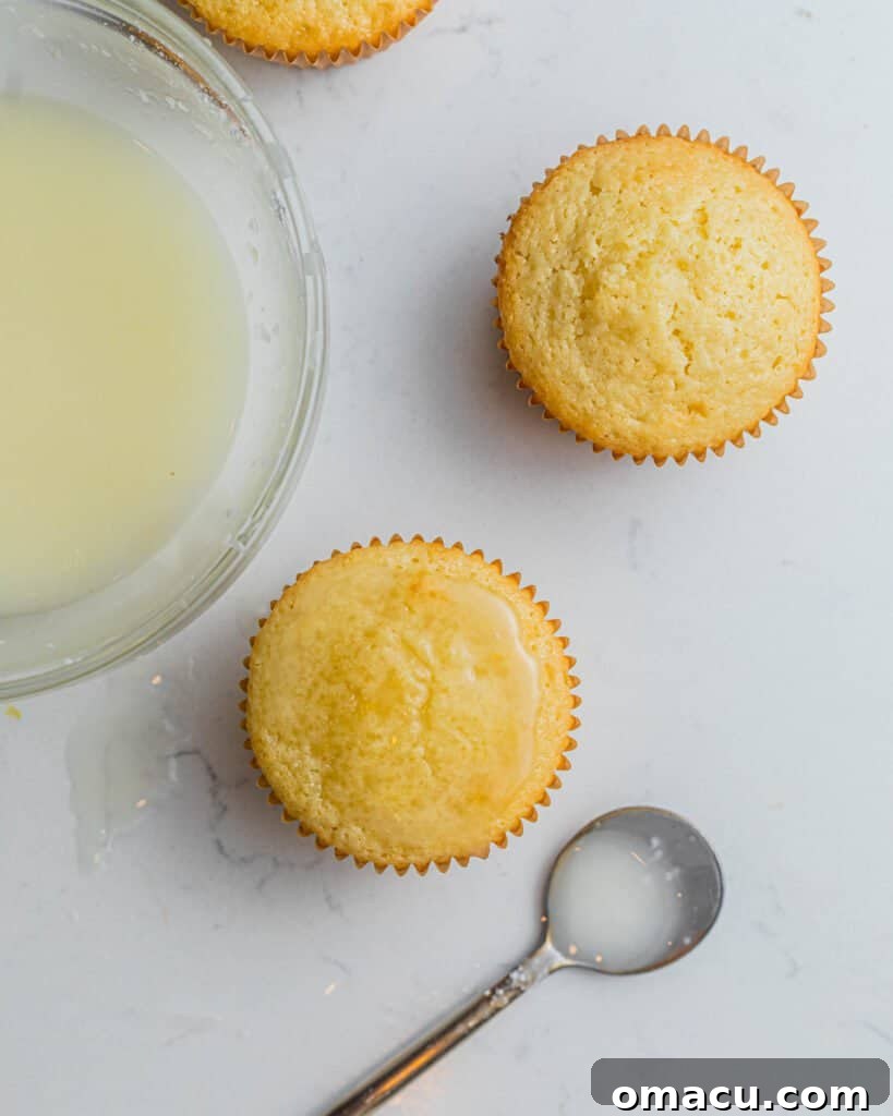 Lemon drizzle being carefully brushed or spooned over the tops of freshly baked cupcakes, showing the glaze soaking into the cake.
