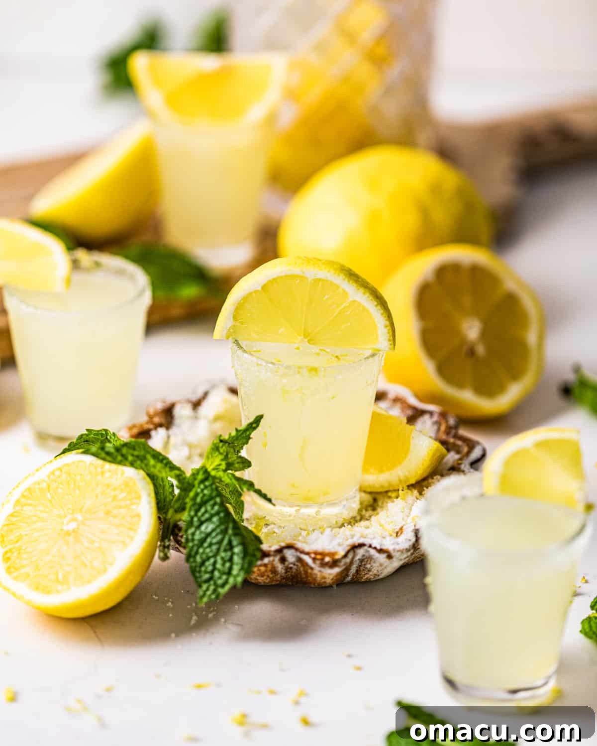 Bright yellow lemon drop shots surrounded by freshly cut lemons and garnished with vibrant mint leaves, set on a dark surface.