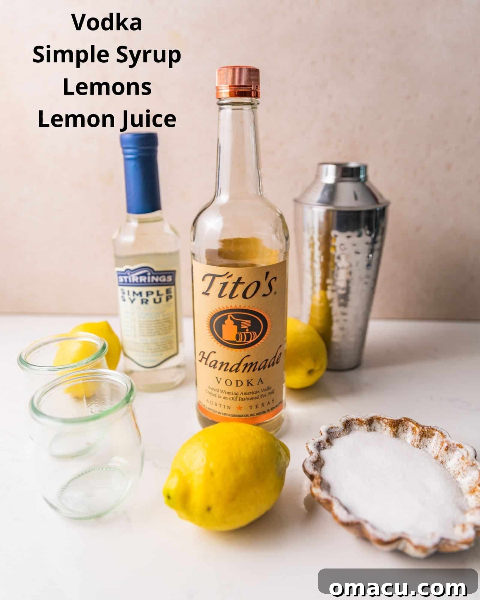 The simple, fresh ingredients required to make a lemon drop shot: a bottle of vodka, a bottle of simple syrup, whole lemons, a container of lemon juice, and a small bowl of white sugar.
