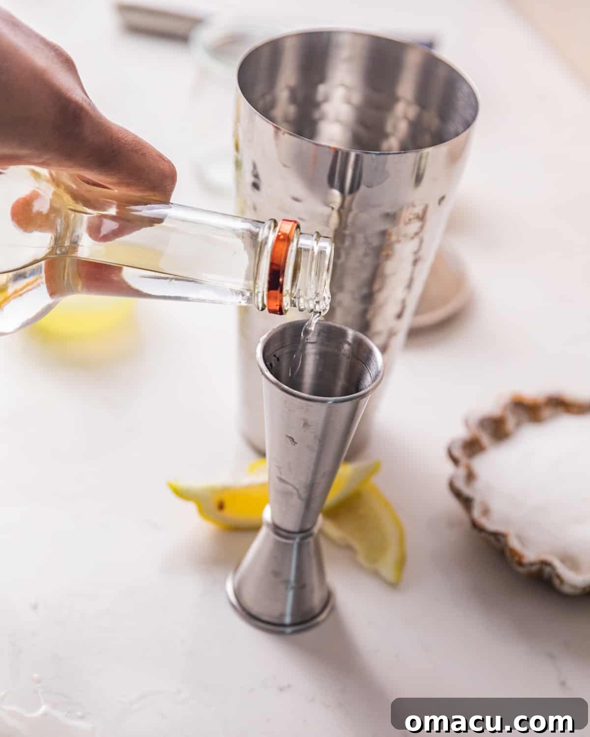 A hand pouring clear vodka from a measuring jigger into a cocktail shaker.