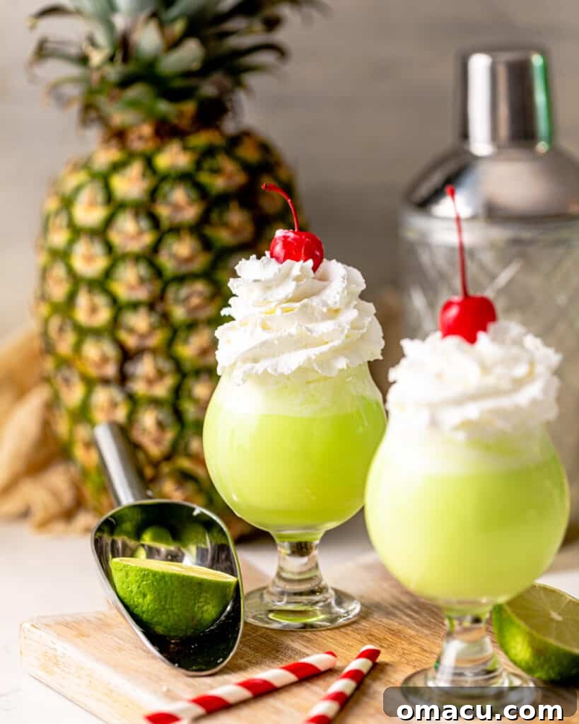 Two vibrant green Scooby Snack Shots garnished with whipped cream and cherries, set against a background of fresh pineapple slices, evoking a tropical vibe.