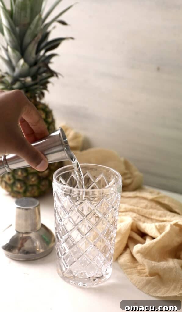 Pouring clear coconut rum into a shiny silver cocktail shaker, with other ingredients waiting nearby.