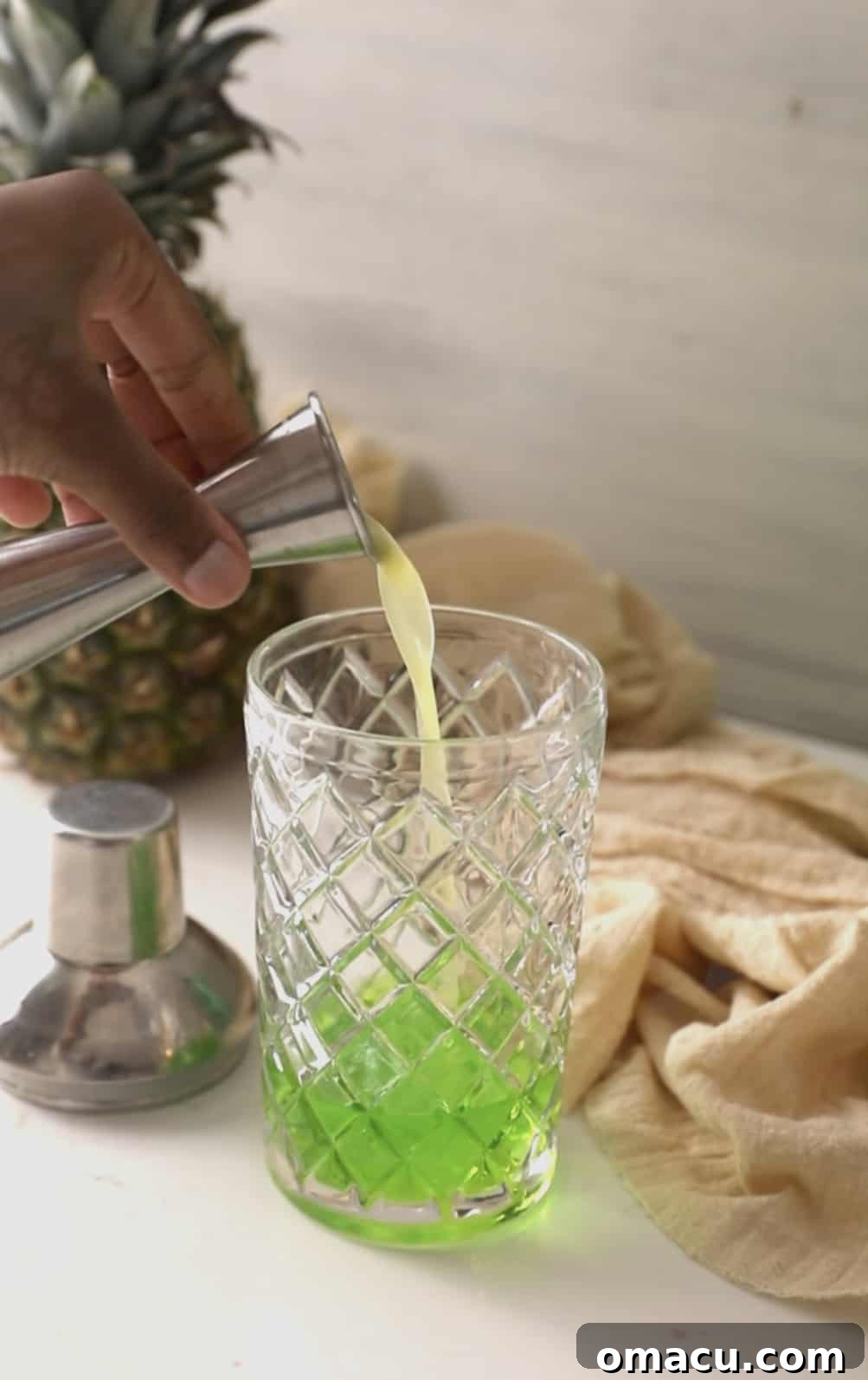 Fresh pineapple juice being poured into the cocktail shaker, adding a layer of yellow to the green and clear liquids.