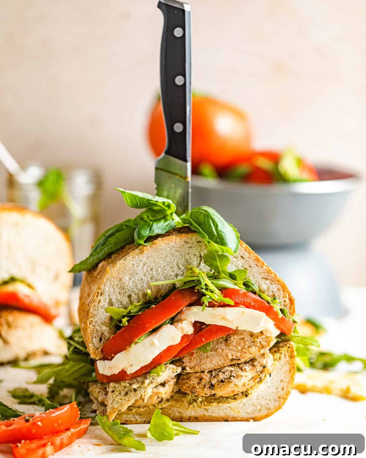 A beautifully sliced Chicken Caprese Sandwich, revealing its fresh layers of chicken, mozzarella, tomatoes, basil, and arugula, positioned in front of a rustic bowl of vibrant red tomatoes.