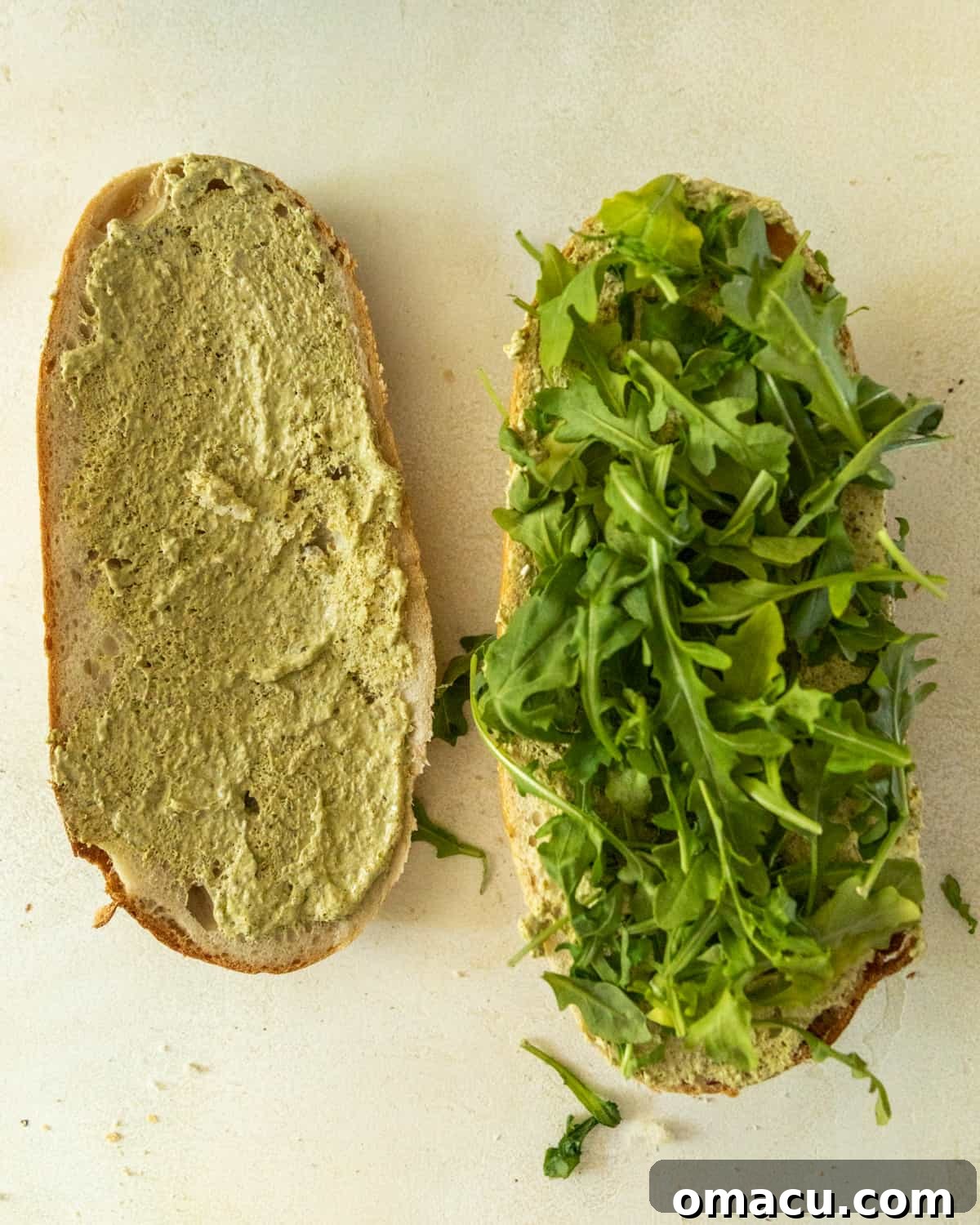 A ciabatta bread loaf sliced in half; one side is generously spread with vibrant pesto mayo, while the other half is topped with fresh arugula, creating a base for the sandwich.