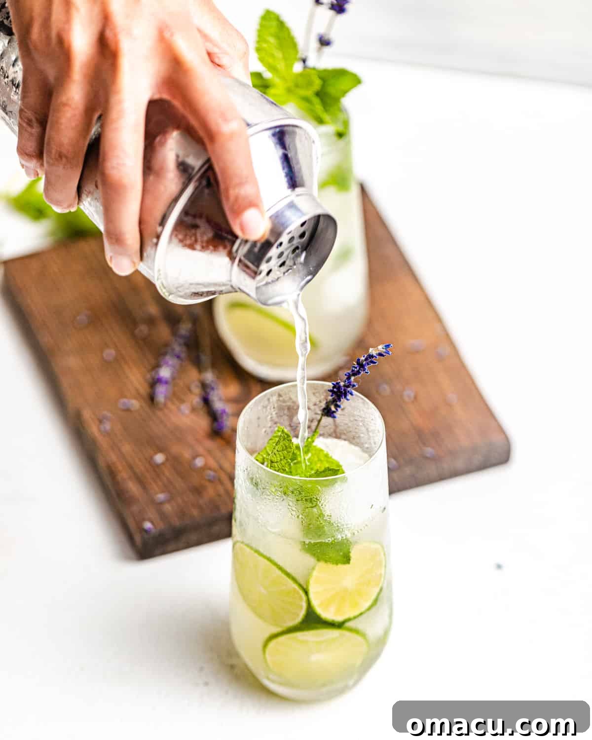 A hand pouring a sparkling gin mojito into a tall highball glass from a cocktail shaker, highlighting the vibrant green mint leaves and lime slices within the drink.