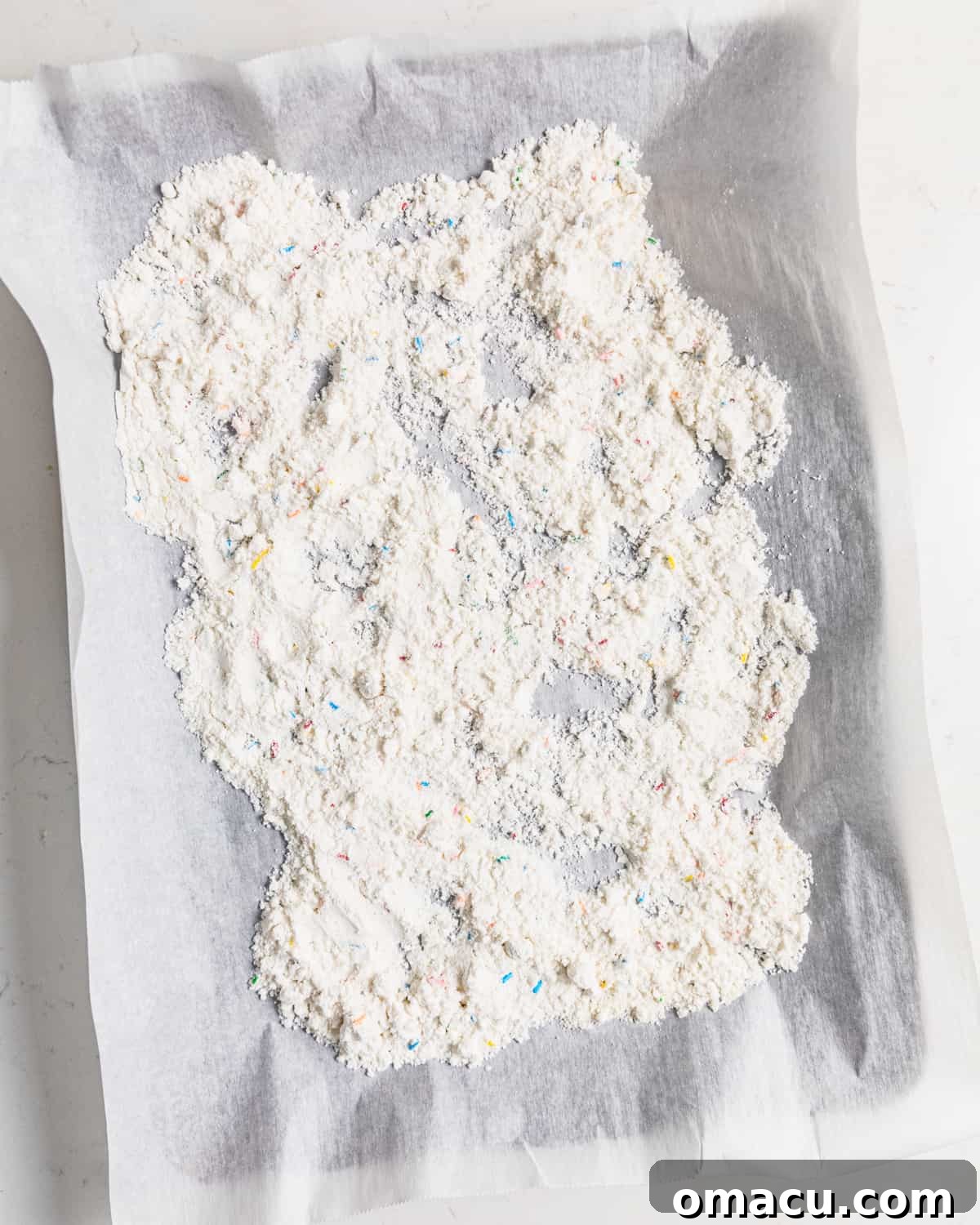 Heat-treated funfetti cake mix spread evenly on a parchment-lined baking sheet, cooling after being baked for safety.