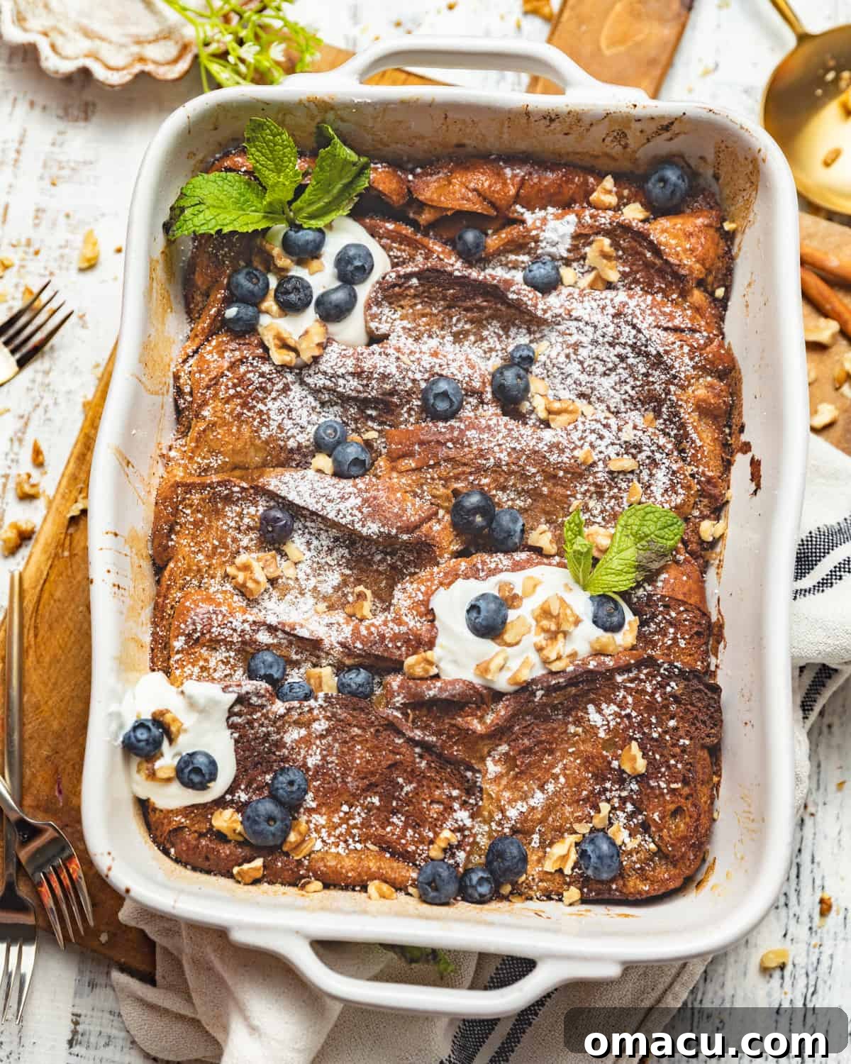 A large brioche French toast casserole in a baking dish, generously topped with fresh blueberries, a swirl of whipped cream, and a sprinkle of chopped walnuts.