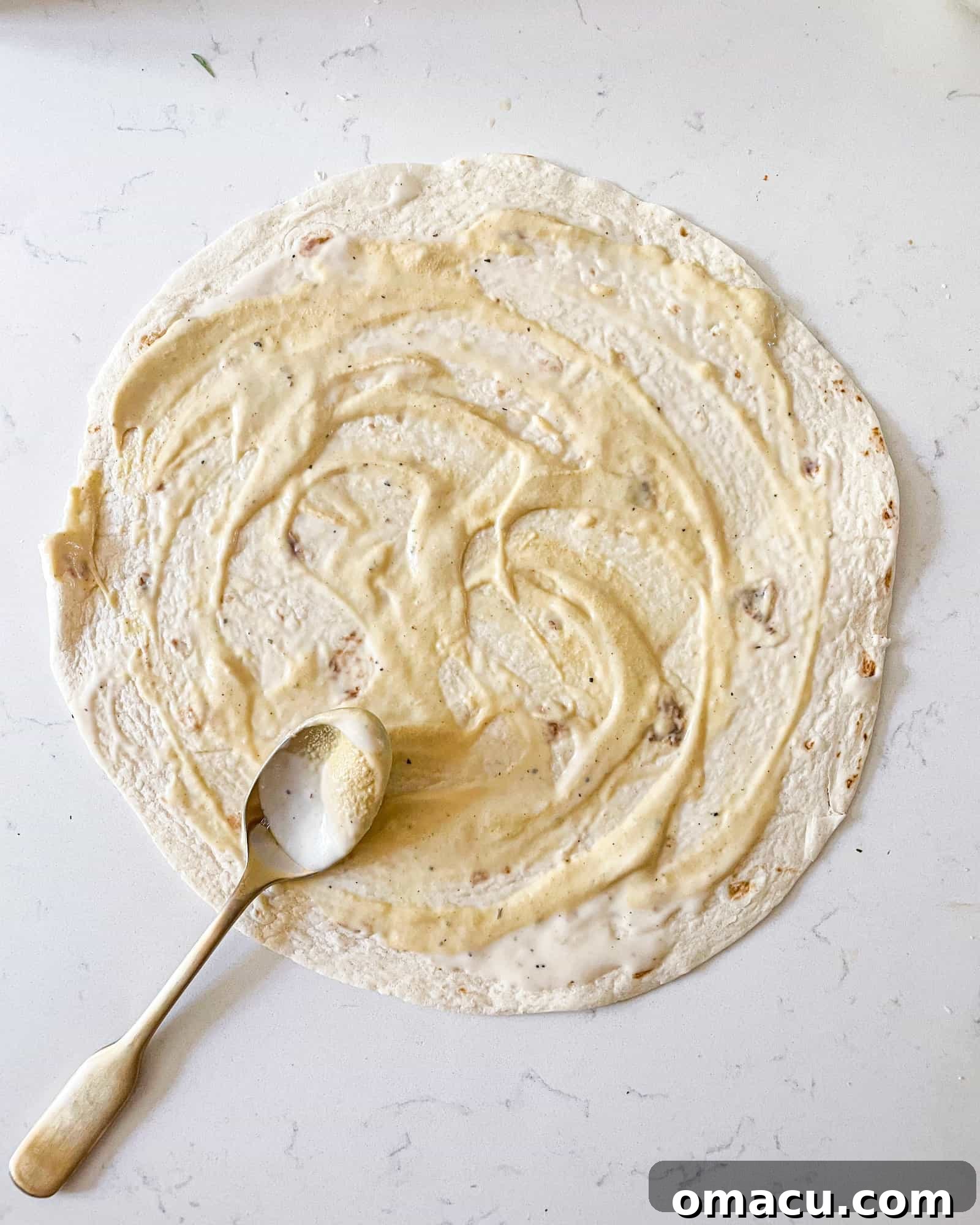 Spreading ranch dressing and dijon mustard on a flour tortilla, the first step in making a chicken bacon ranch wrap.