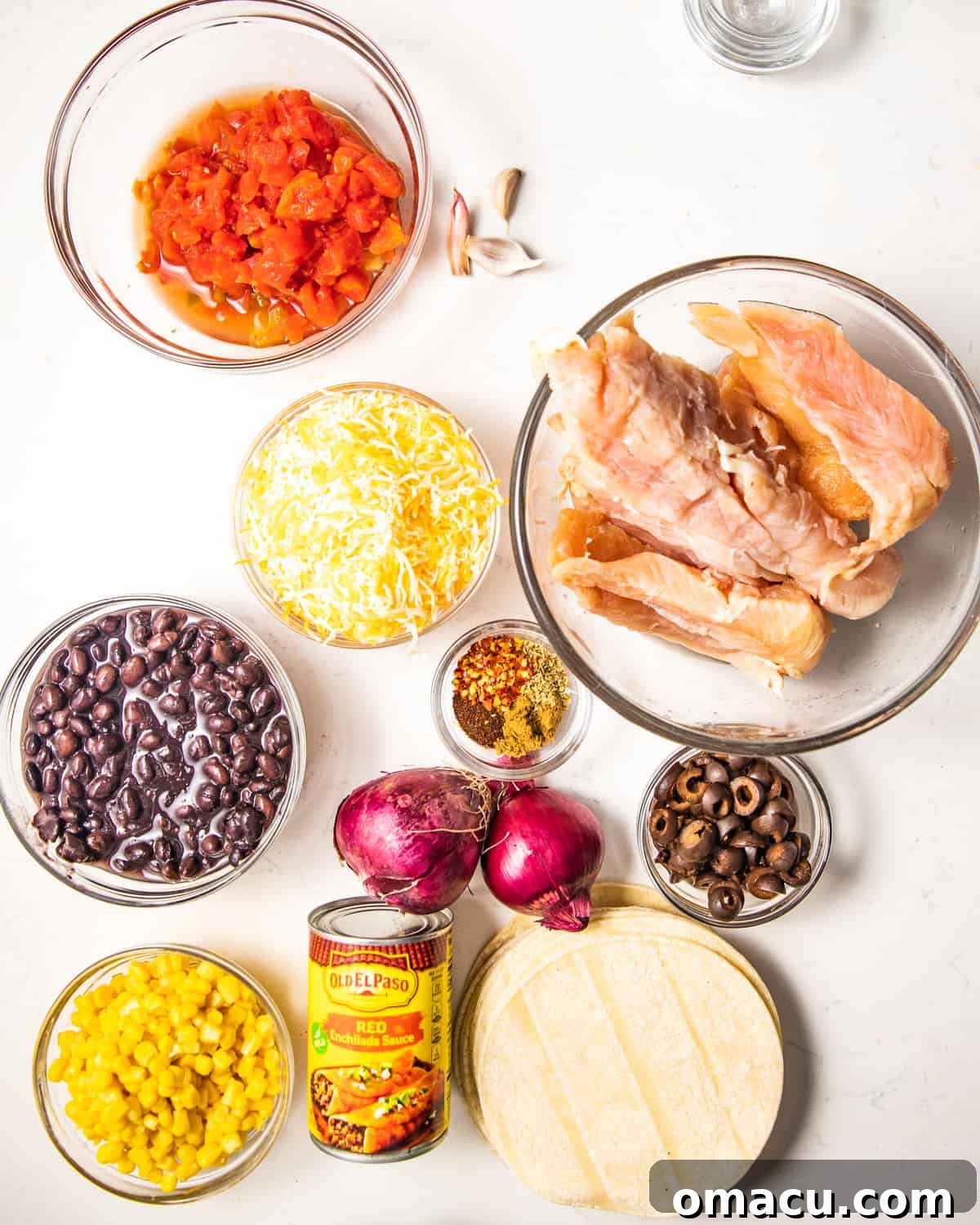 A vibrant selection of ingredients laid out for making chicken enchiladas, including ripe tomatoes, raw chicken breasts, sliced red onion, canned black beans, sweet corn, various seasonings, soft tortillas, rich enchilada sauce, black olives, and shredded cheese.