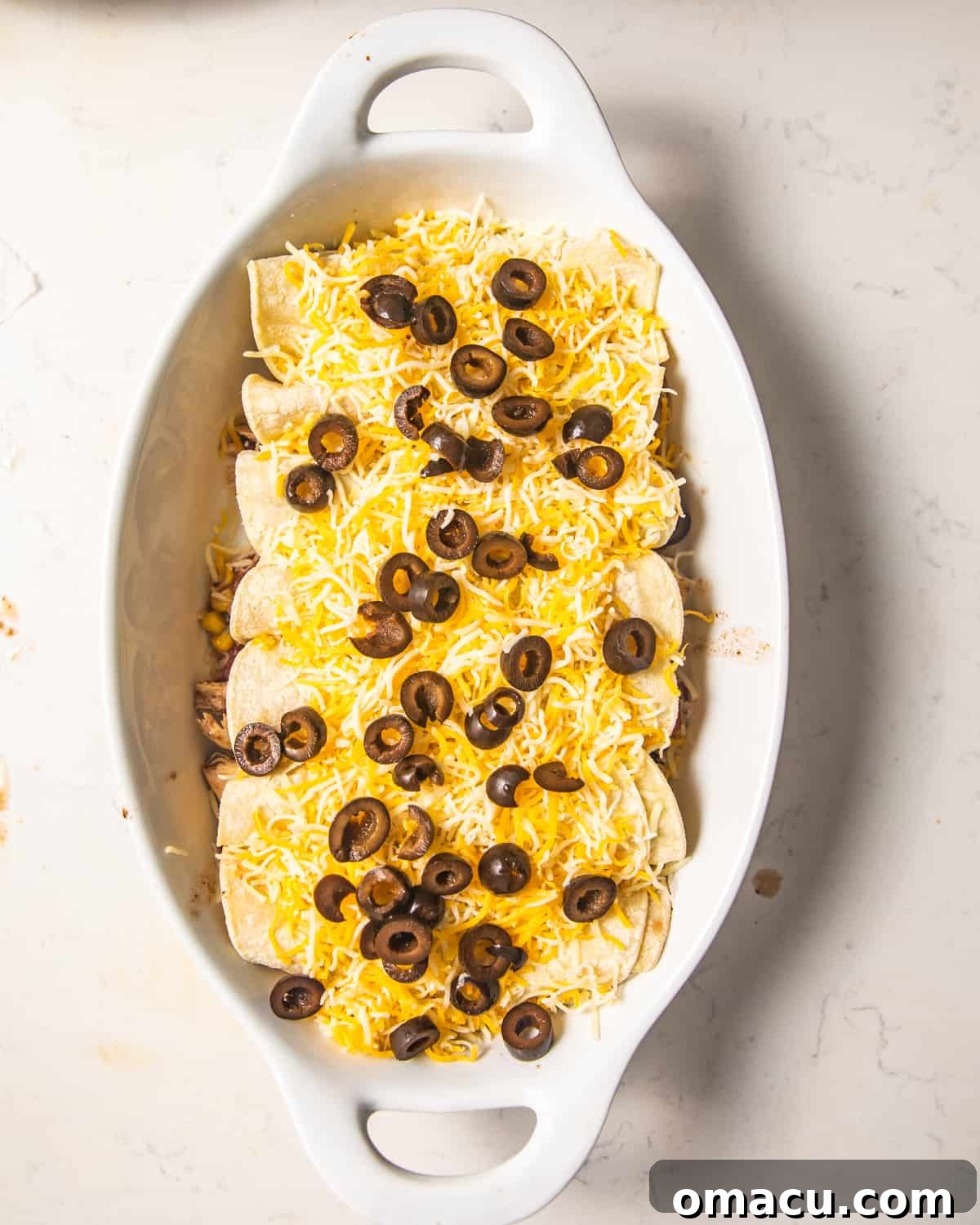 Assembled chicken enchiladas in a casserole dish, generously topped with shredded cheese and black olives, poised for baking.