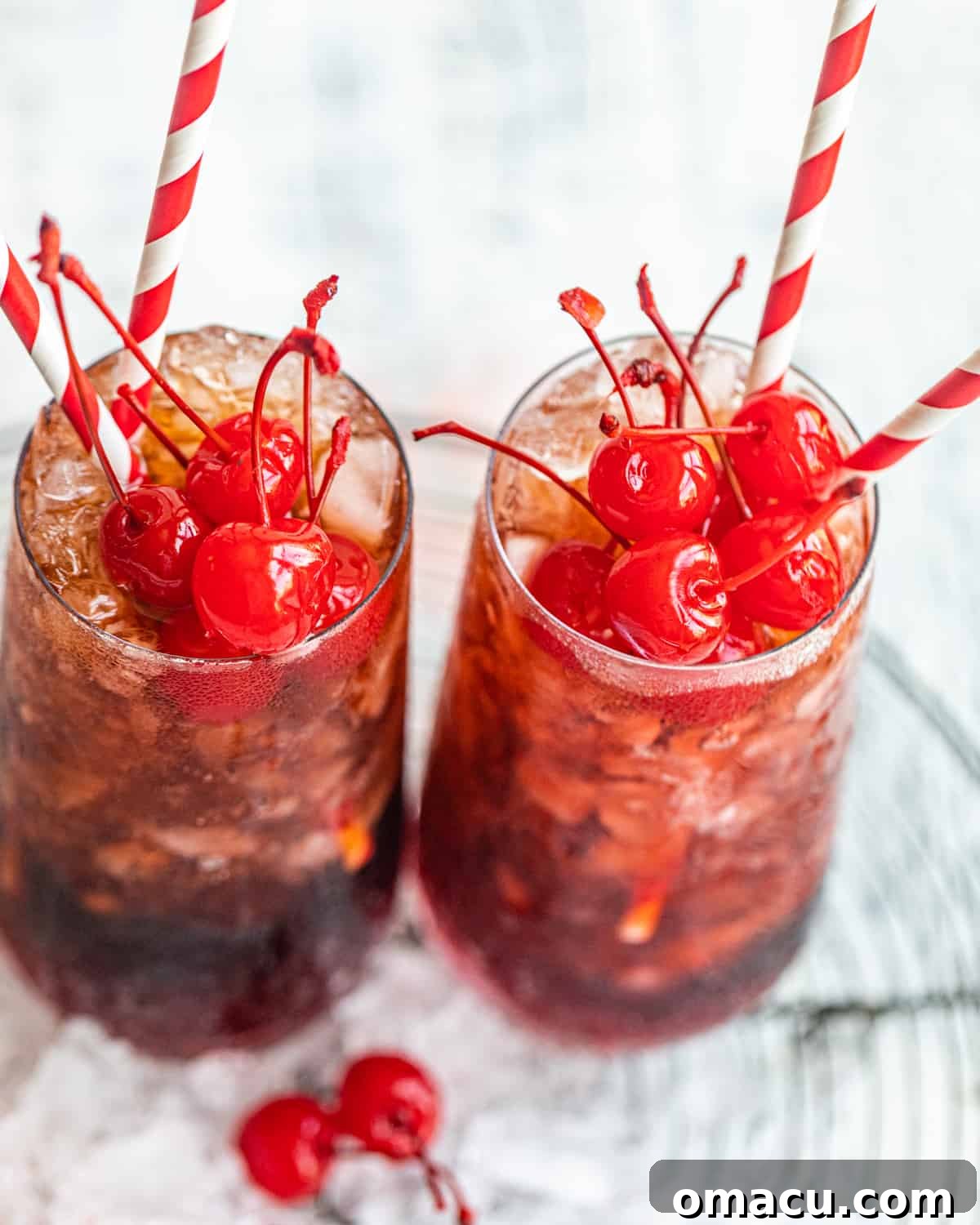 Two vibrant Roy Rogers drinks displayed on a wire rack, adorned with ice cubes and bright maraschino cherries, ready to be served.