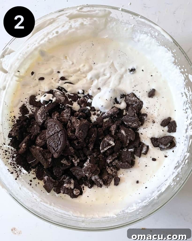 Crushed Oreo cookies being folded into the creamy vanilla ice cream mixture in a bowl, creating the classic cookies and cream swirl.