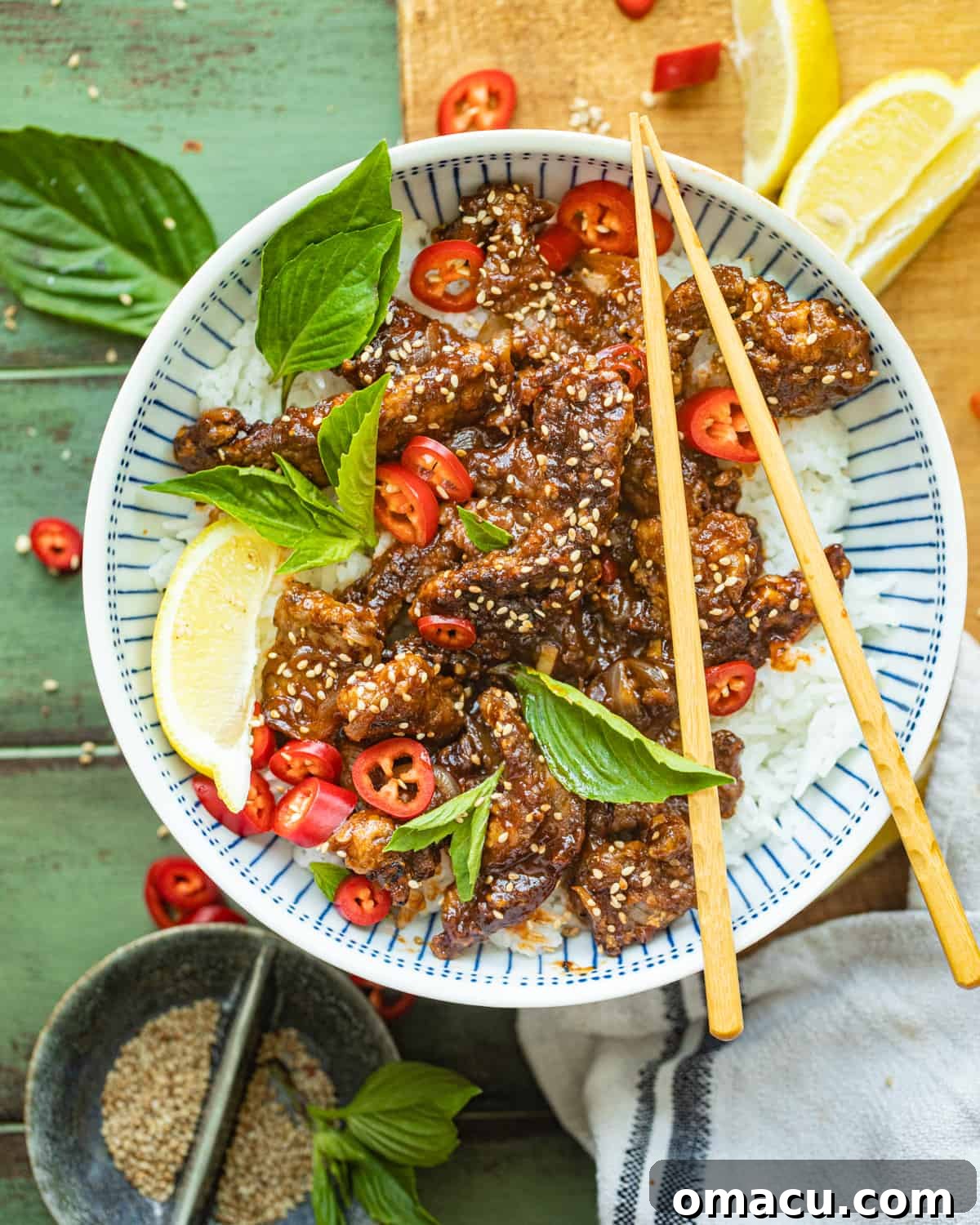 A bowl of crispy chilli beef, garnished with fresh herbs and sesame seeds, with chopsticks ready for serving.