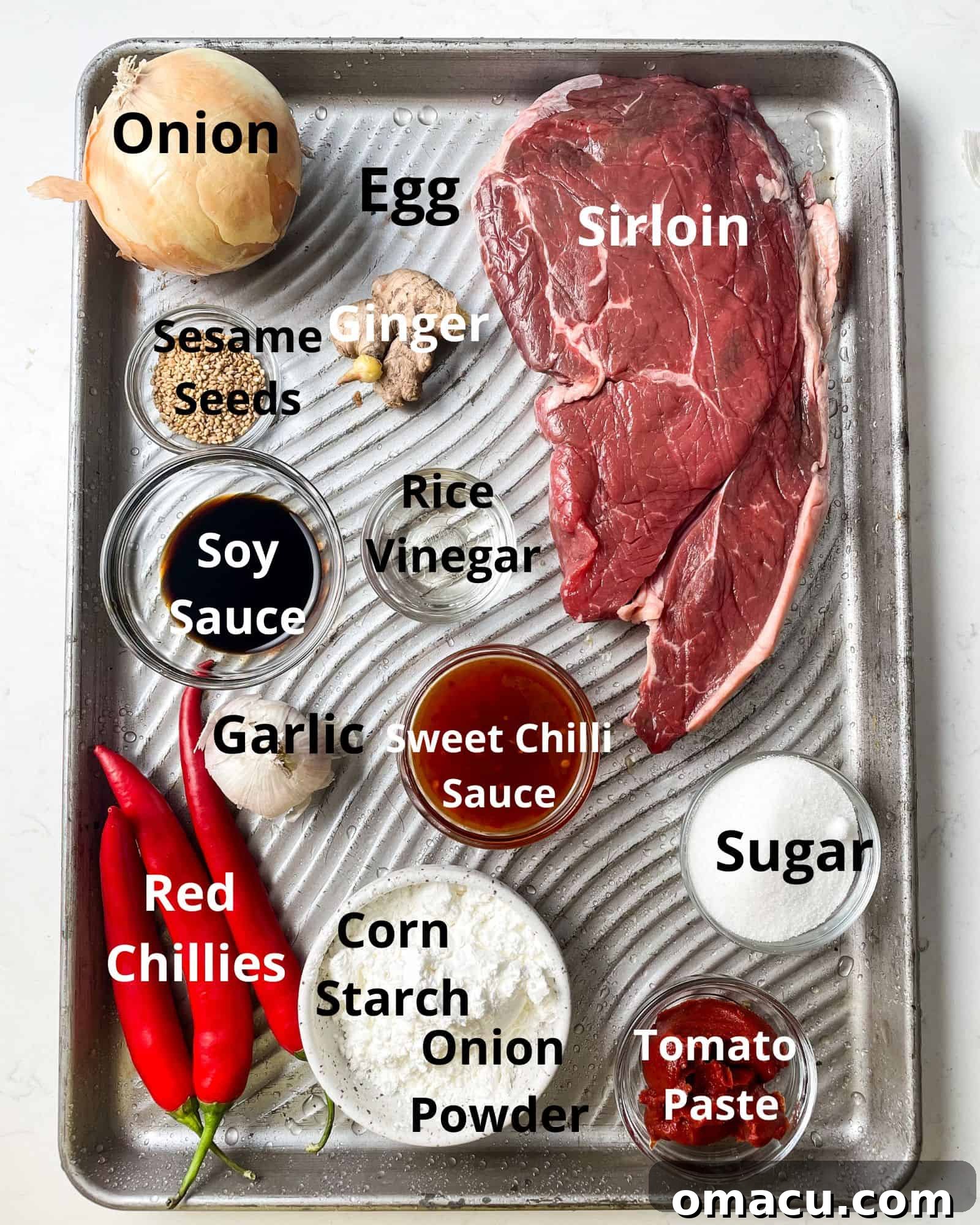 A selection of fresh and pantry ingredients laid out for making crispy chilli beef: sirloin, egg, corn starch, salt, pepper, onion powder, garlic cloves, granulated sugar, vibrant red chillies, soy sauce, a sliced onion, white sesame seeds, rich tomato paste, and a bottle of sweet chilli sauce, alongside rice vinegar.
