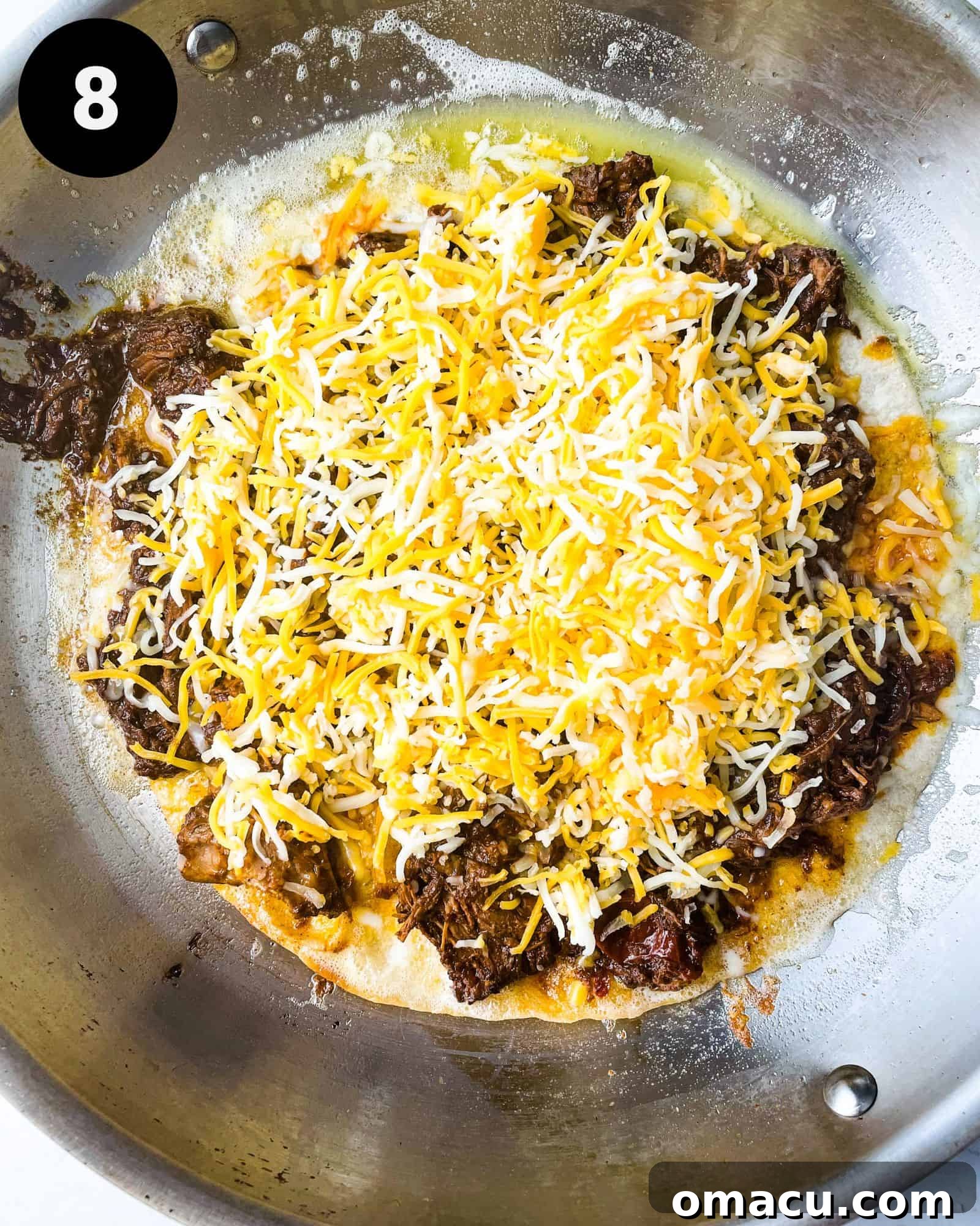 a tortilla in a pan topped with birria and cheese, awaiting the second tortilla.