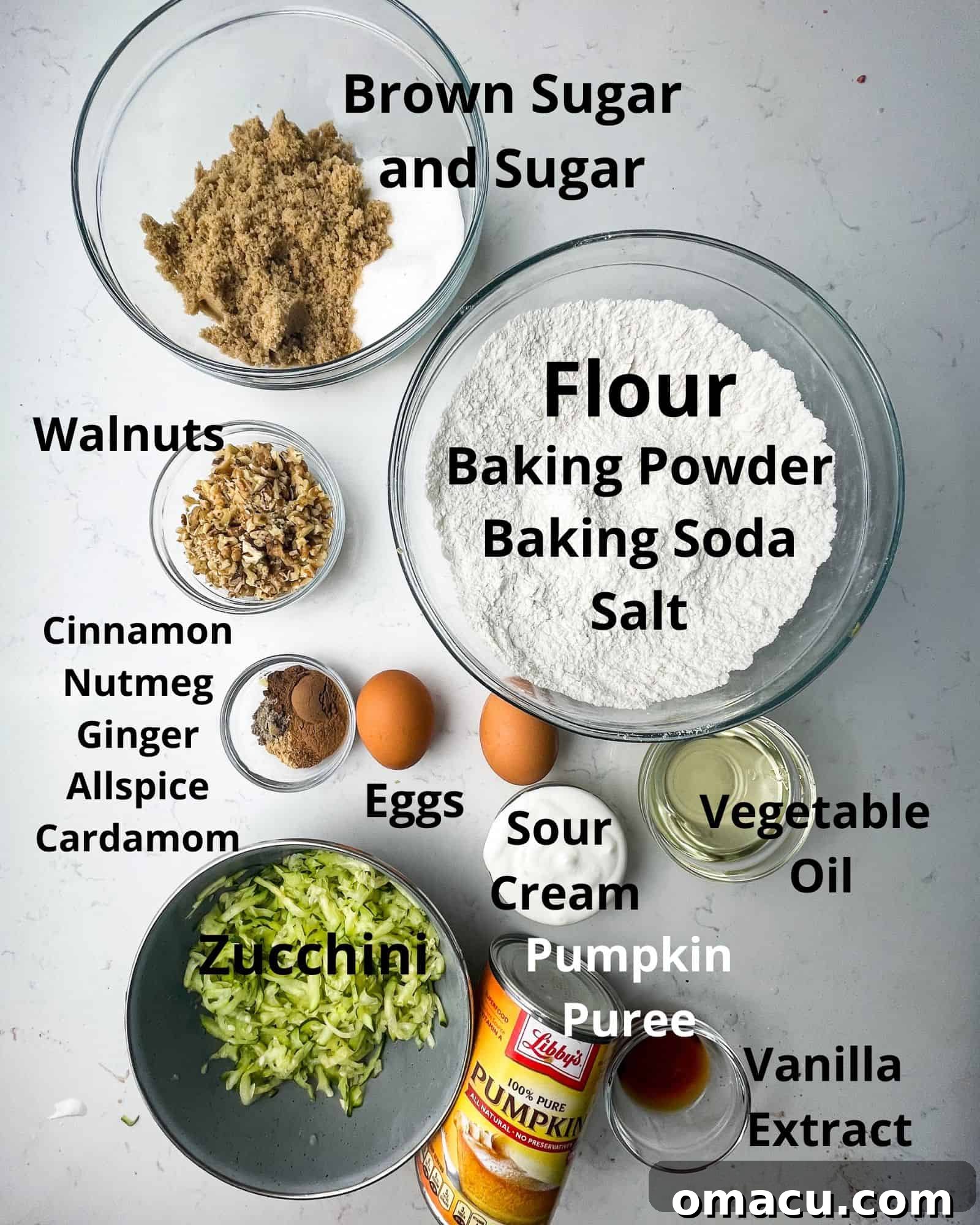 A colorful array of ingredients for zucchini pumpkin muffins laid out on a clean surface: flour, baking soda, baking powder, salt, a variety of pumpkin spices, brown sugar, white sugar, vegetable oil, sour cream, eggs, vanilla extract, pumpkin puree, fresh zucchini, and walnuts.