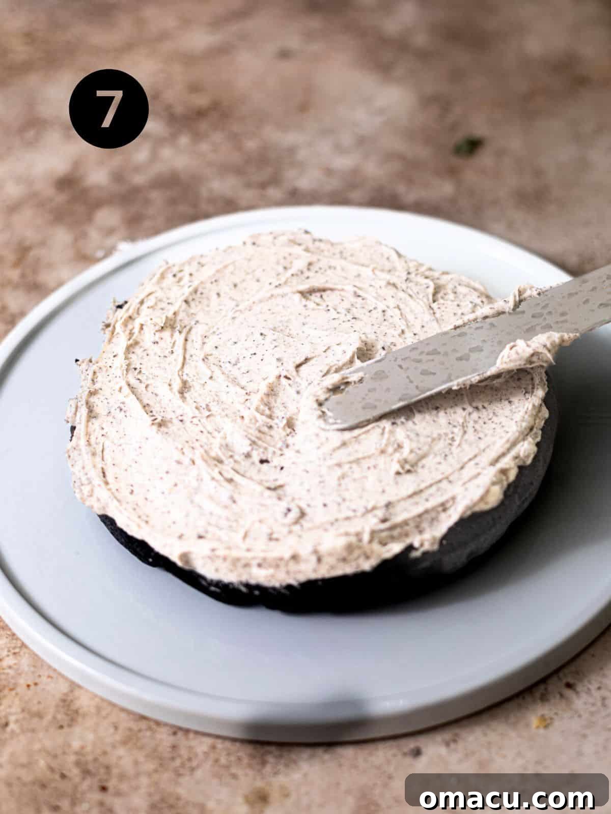A layer of Oreo frosting being spread evenly onto a dark black velvet cake layer on a cake stand, with subsequent layers visible.