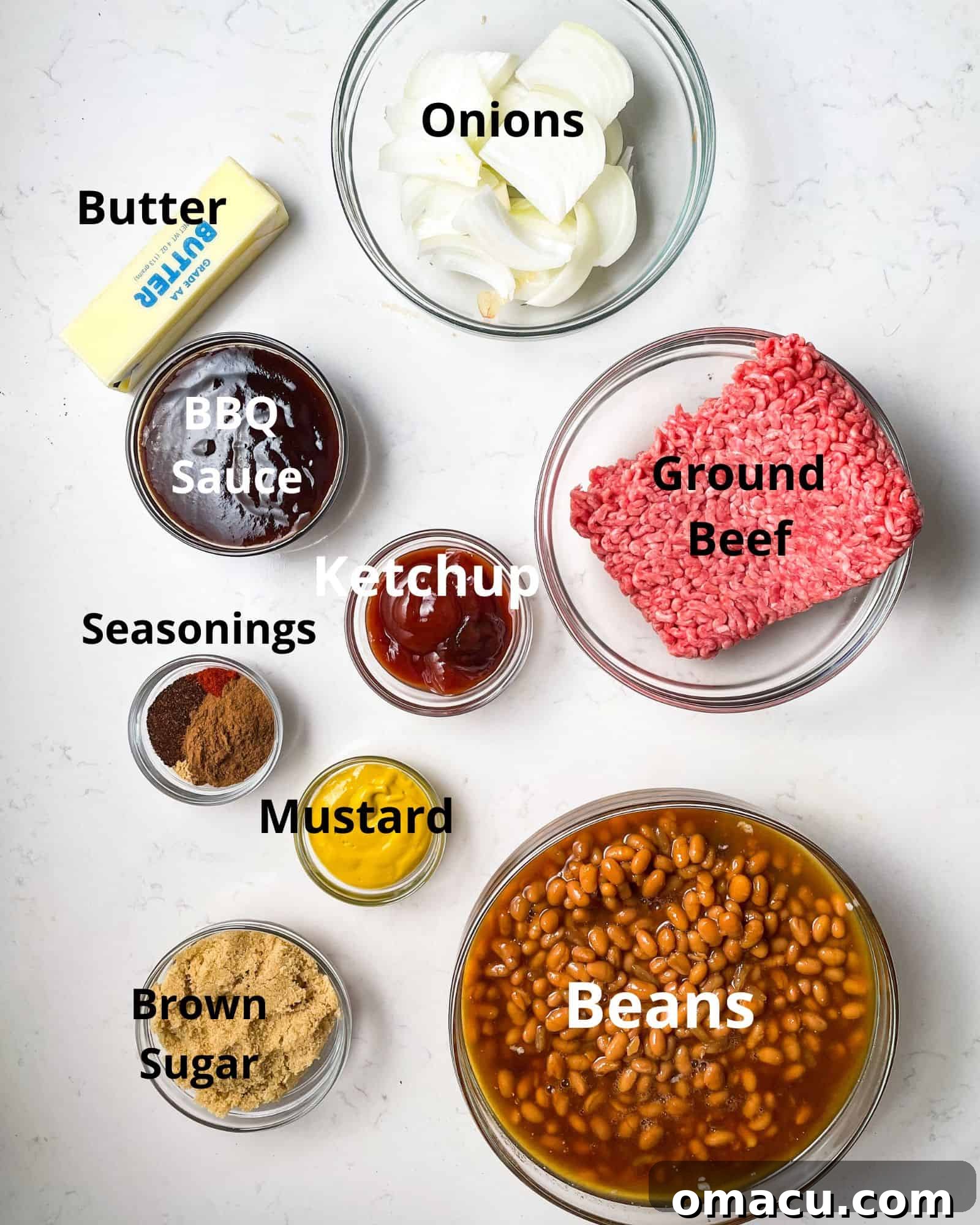ingredients to make baked beans - canned beans, ground beef, onions, butter, bbq sauce, ketchup, mustard, seasonings, and brown sugar laid out on a wooden surface.