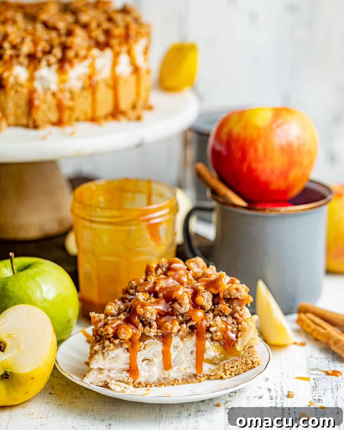 A perfectly sliced piece of Apple Pie Stuffed Cheesecake on a pristine white plate, garnished with a sweet caramel drizzle, with a subtly blurred cup in the background.