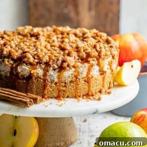 A vibrant close-up of the Apple Pie Stuffed Cheesecake on a cake stand, with the crumble topping and caramel drizzle visible.