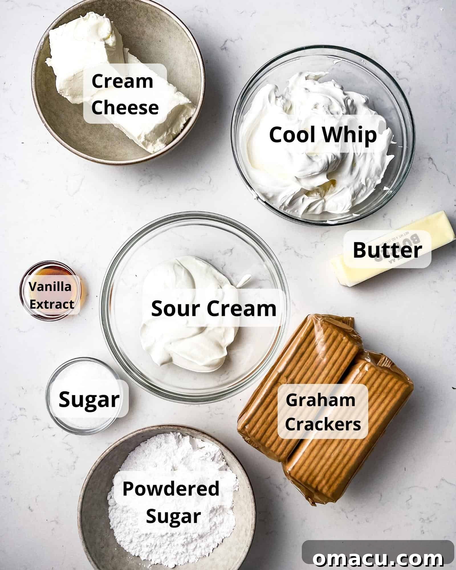 All the essential ingredients neatly arranged to make Philadelphia No-Bake Cheesecake: cream cheese, cool whip, sour cream, butter, vanilla extract, graham crackers, powdered sugar, and granulated sugar.