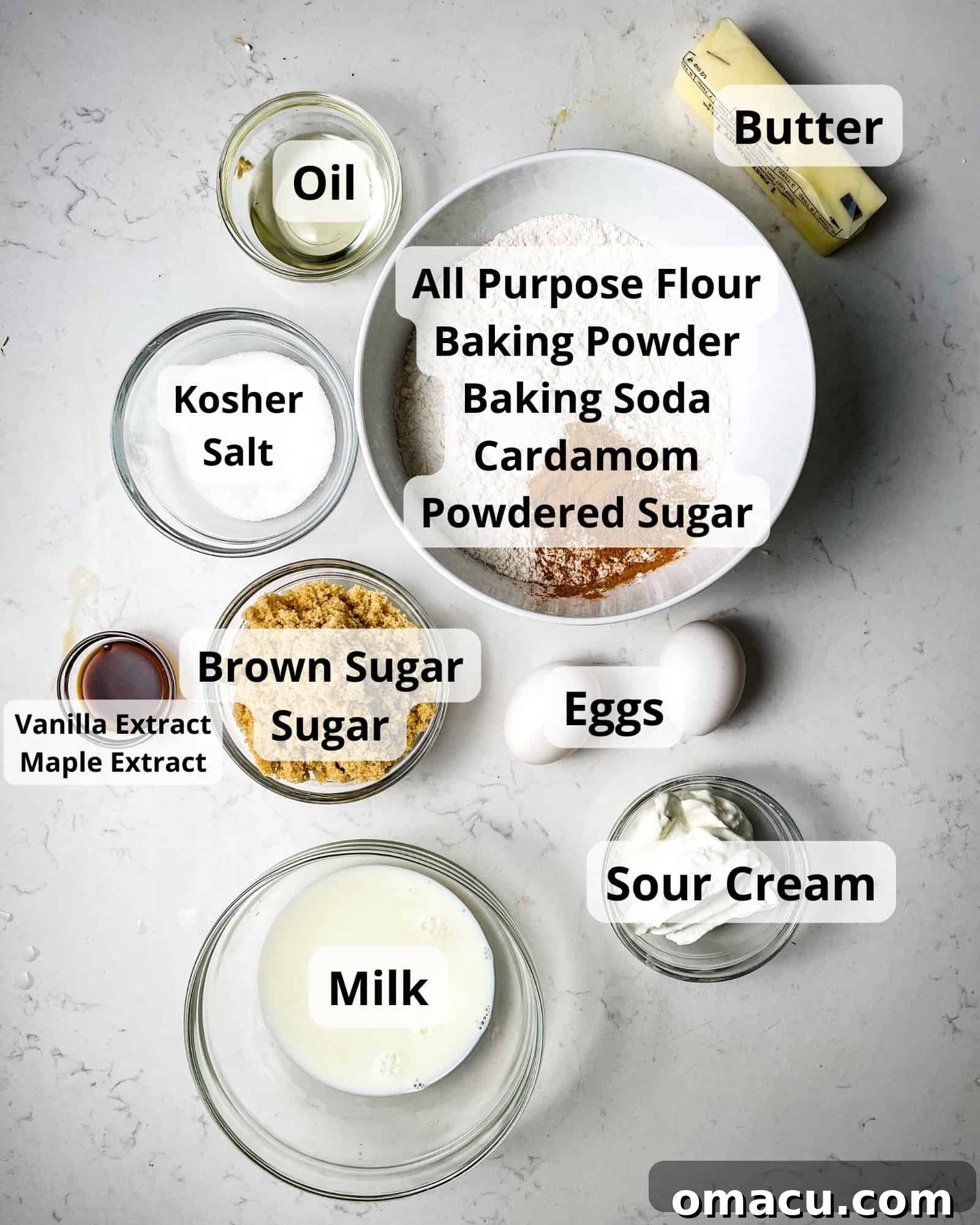 Arrangement of ingredients for cardamom cake: flour, sugar, brown sugar, sour cream, oil, eggs, milk, cardamom, baking powder, baking soda, butter, and vanilla extract.