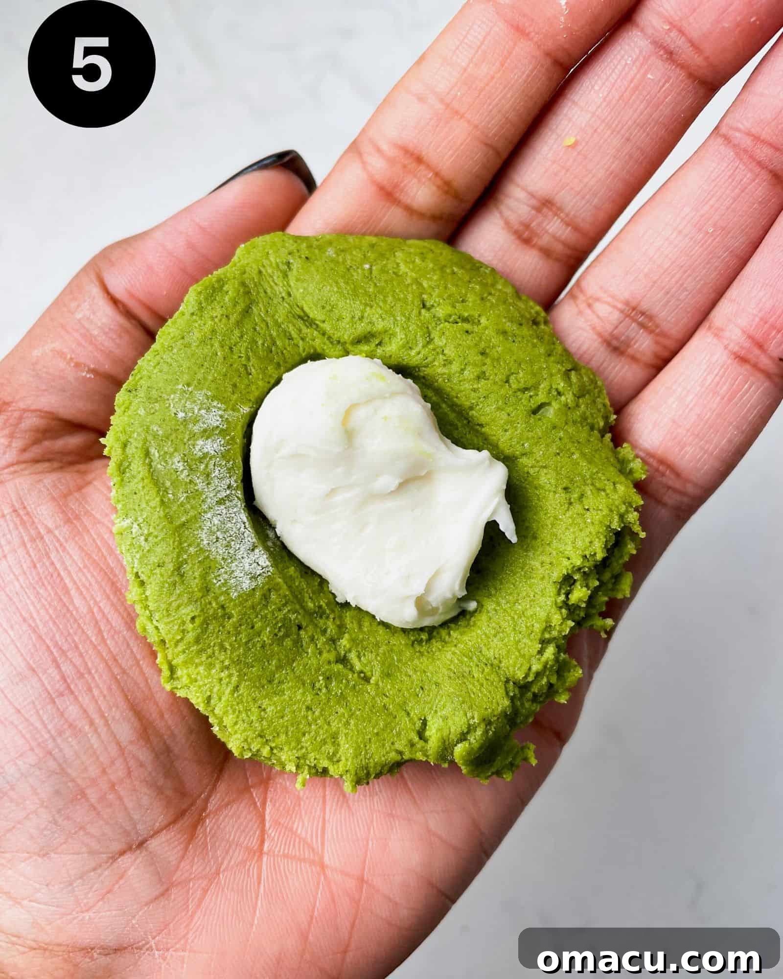 A matcha cookie dough ball, gently pressed open to reveal a luscious cream cheese and white chocolate filling nestled inside, ready to be sealed.
