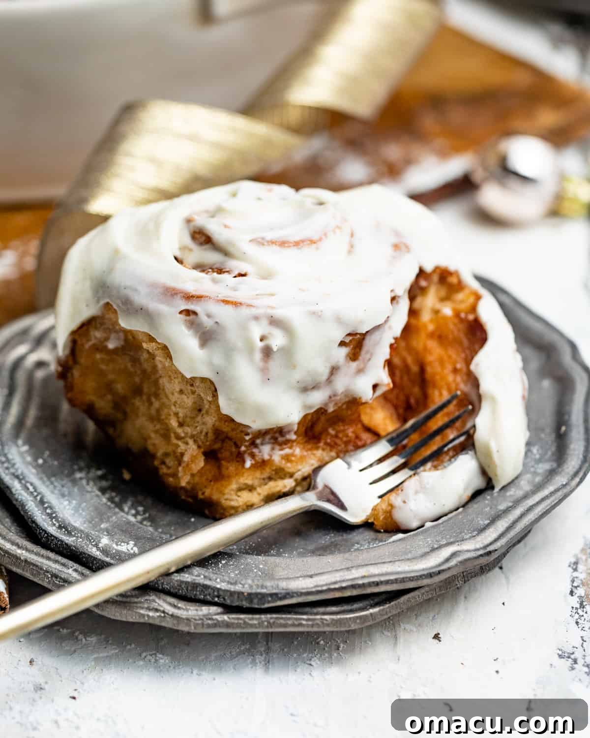 A single, perfectly gooey cinnamon bun on a white plate with a fork, showing off its rich frosting and swirl.