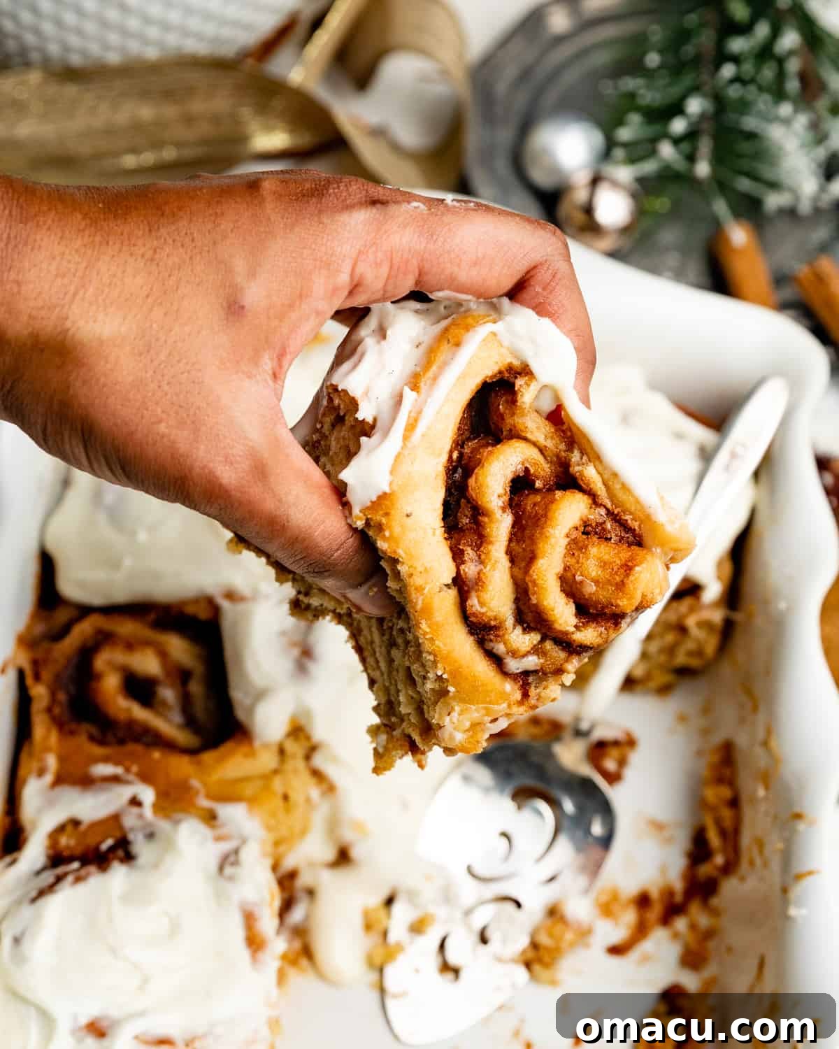 A hand reaching into a baking dish to pick up a warm, frosted cinnamon roll, highlighting its perfect texture.