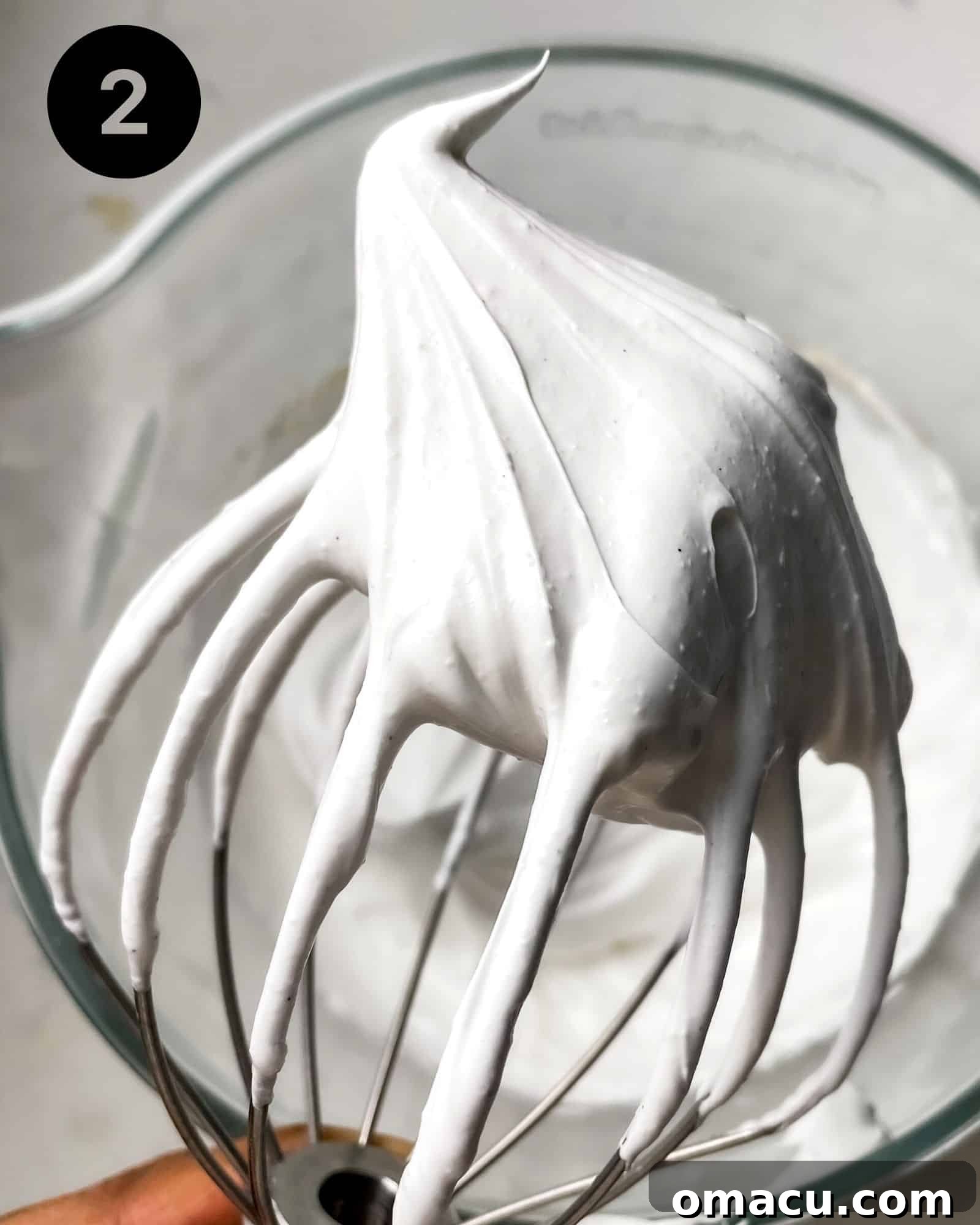 meringue on a whisk showing stiff peaks. A whisk lifted from a bowl, showcasing perfectly formed, stiff, and glossy meringue peaks, indicating readiness for piping.