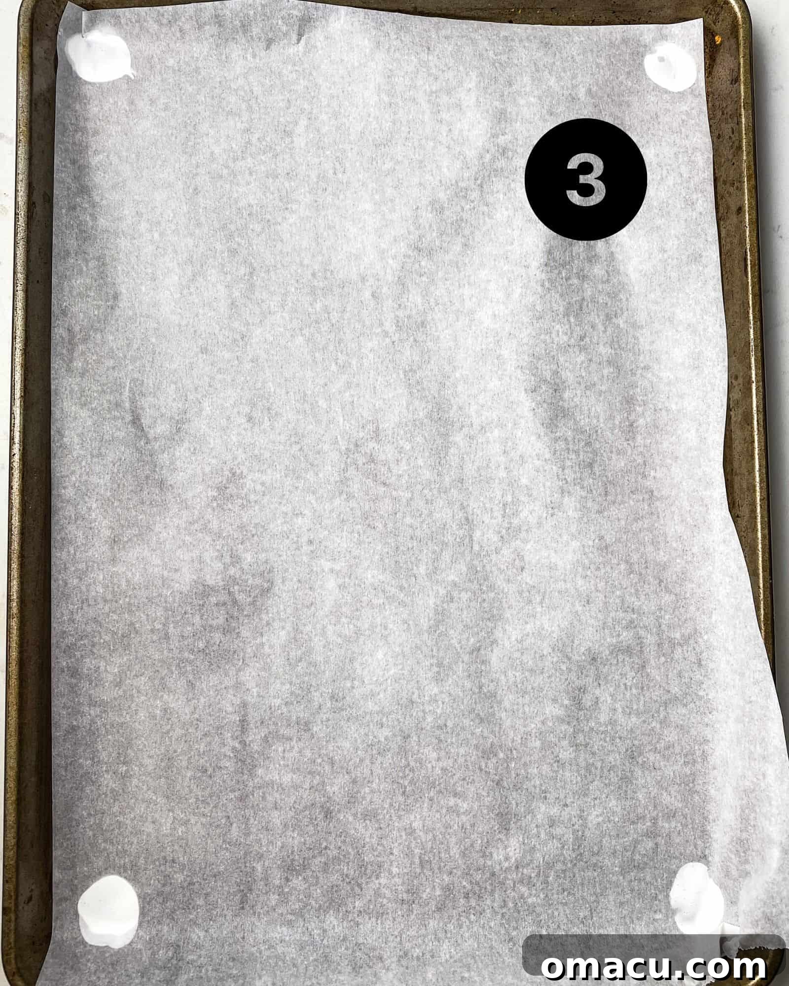 parchment paper on a baking sheet with meringue in four corners to keep the parchment paper from lifting up. A baking sheet lined with parchment paper, secured by small dabs of meringue in each corner, demonstrating a technique to prevent the paper from shifting while piping.