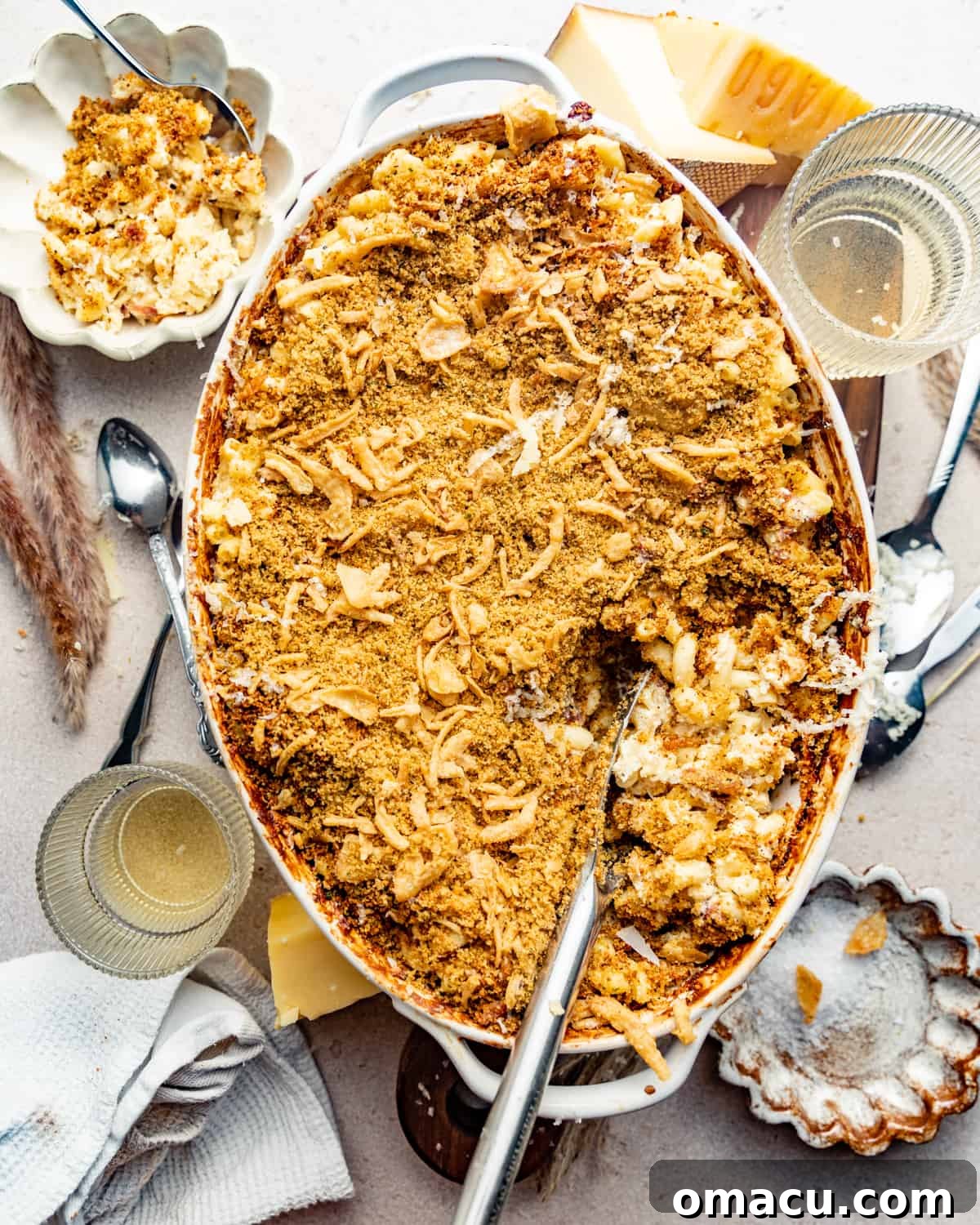 A lavish baking dish of golden-brown prosciutto mac and cheese, artfully surrounded by blocks of cheese, elegant serving spoons, and glasses ready for a feast. The crispy topping is visible.