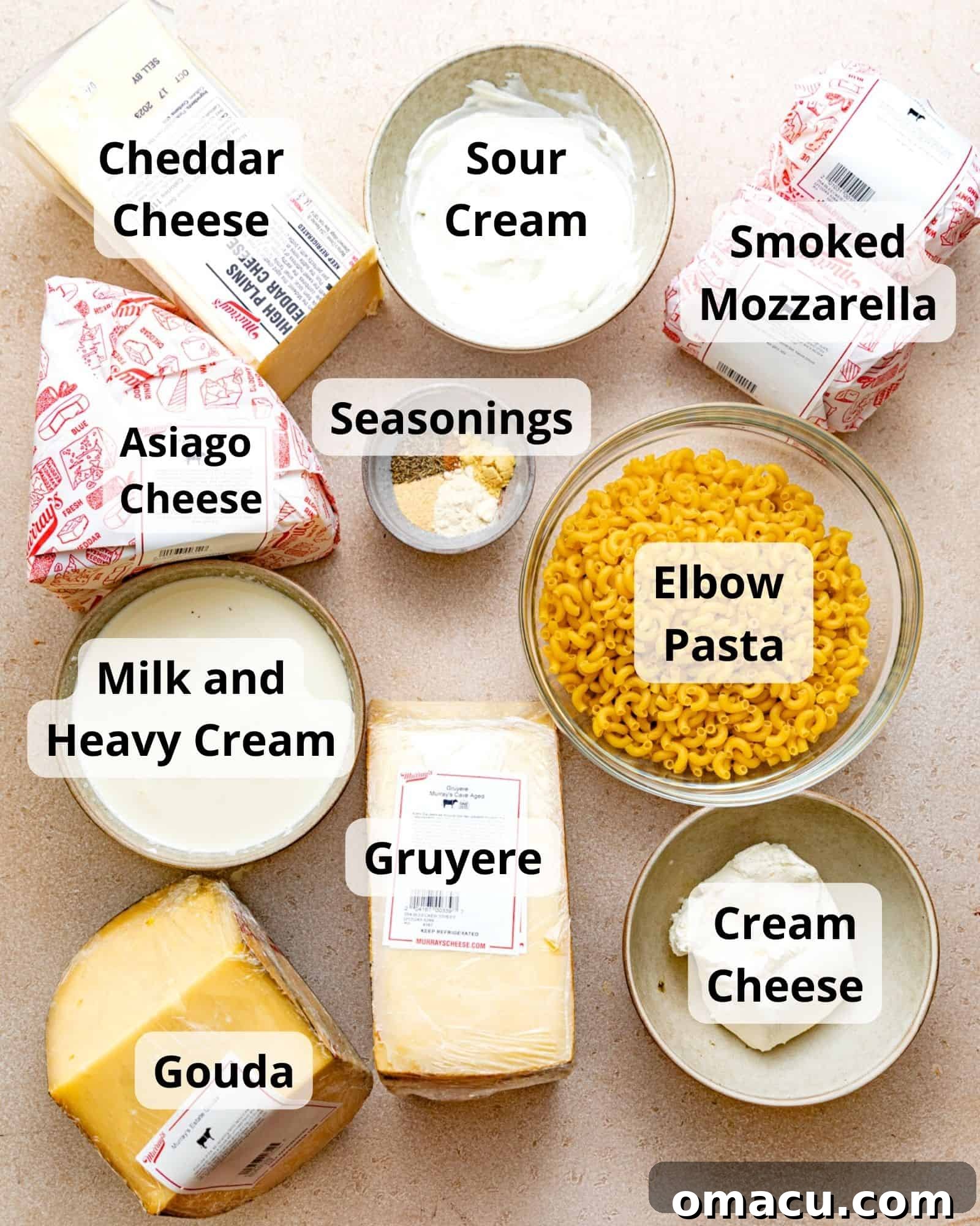 A vibrant display of fresh ingredients laid out for making mac and cheese, including various blocks of cheddar, asiago, smoked mozzarella, gouda, and gruyere cheeses, alongside elbow pasta, sour cream, milk, heavy cream, cream cheese, and a selection of aromatic seasonings.