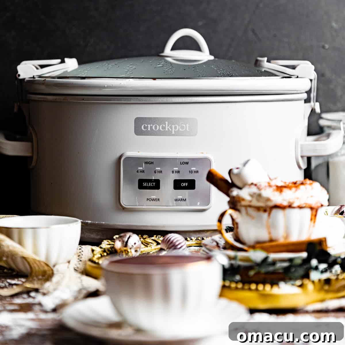 A crockpot slow cooker in the background with cups of hot chocolate in the foreground, ready to serve.