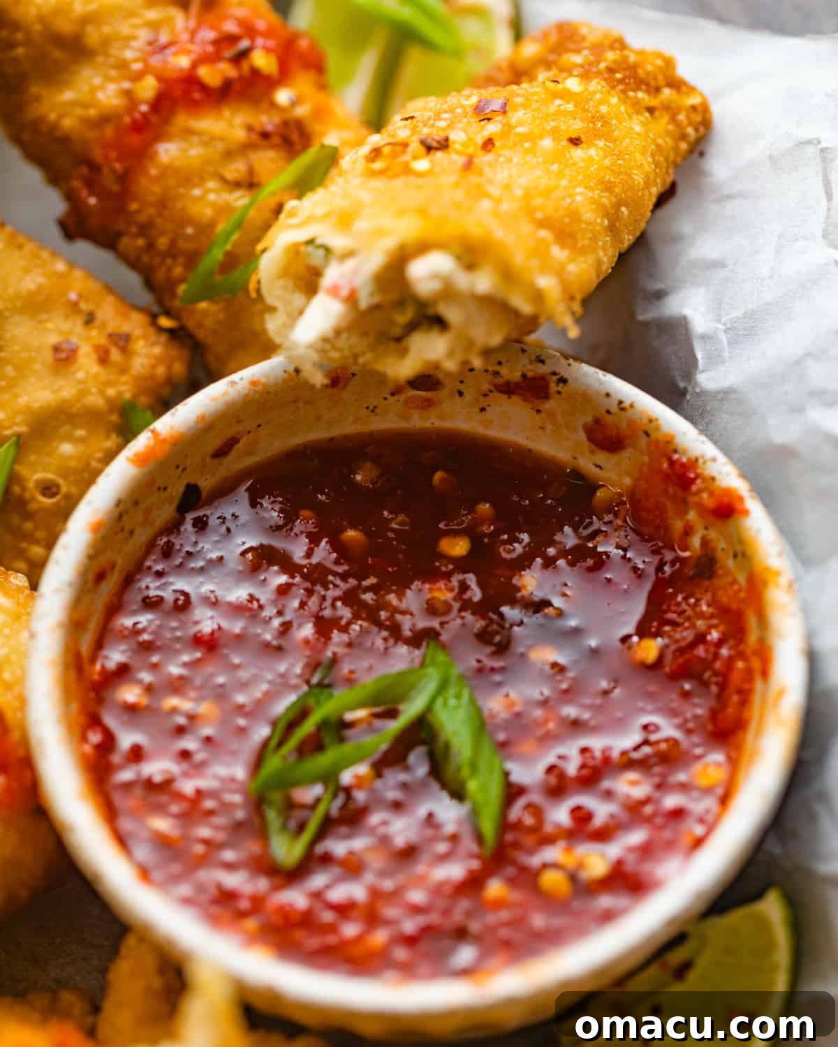 A close-up shot of a bowl filled with vibrant red spicy sweet and sour sauce, with a perfectly fried crab rangoon egg roll resting on top, garnished with red pepper flakes.