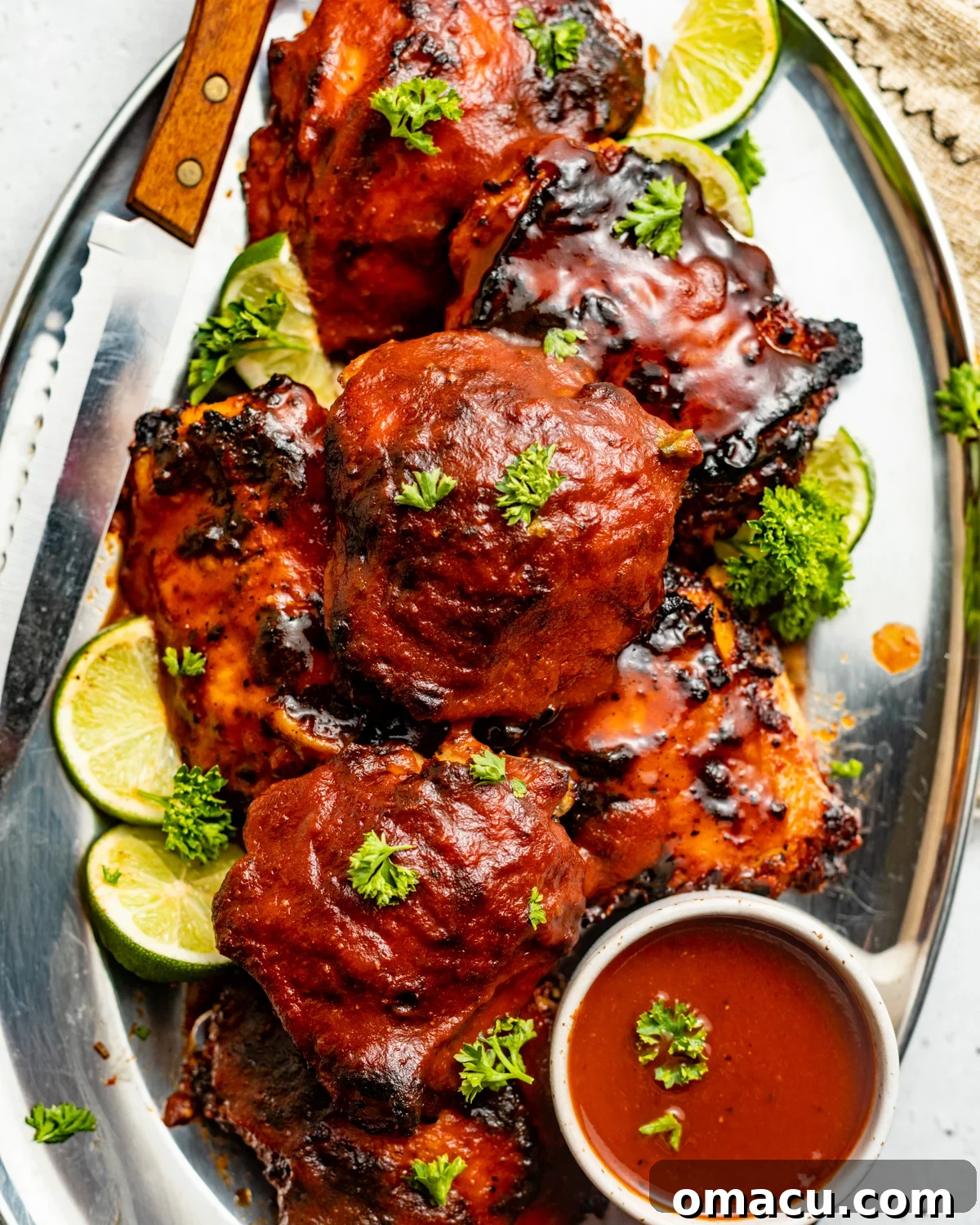 Two baked bbq chicken thighs on a white plate, garnished with fresh herbs, alongside a small bowl of extra bbq sauce and a knife.