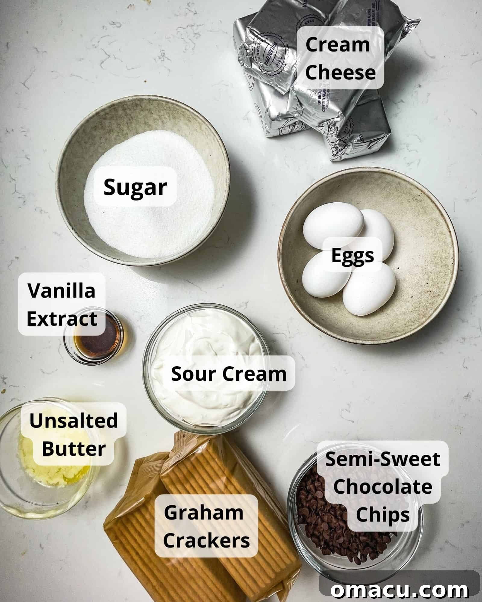 A carefully arranged collection of ingredients for chocolate chip cheesecake, including cream cheese, sugar, eggs, vanilla extract, sour cream, melted butter, graham crackers, chocolate chips, and heavy cream.