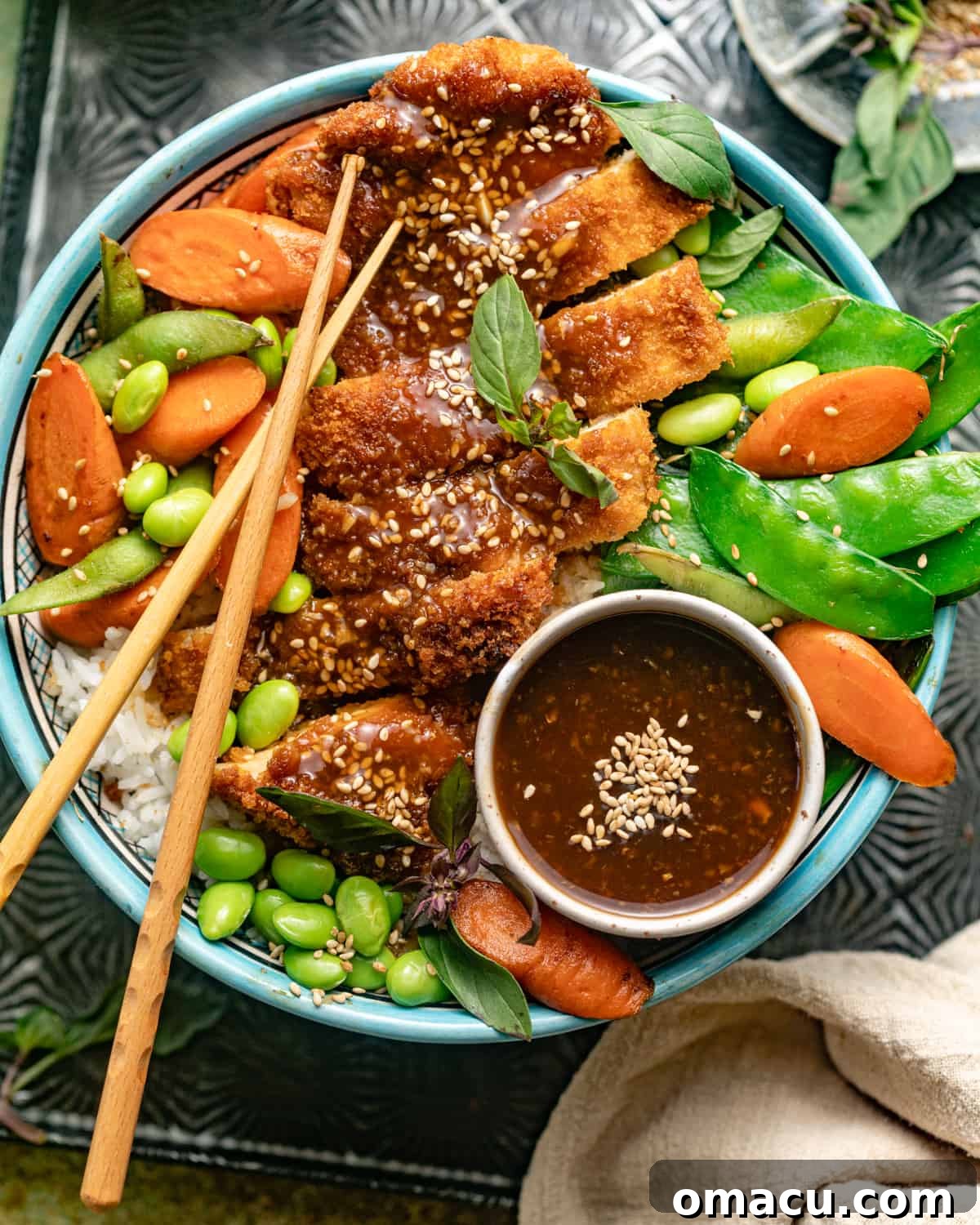 Teriyaki chicken, edamame beans, carrots, snow peas, and rice in a large bowl with chopsticks and teriyaki sauce.