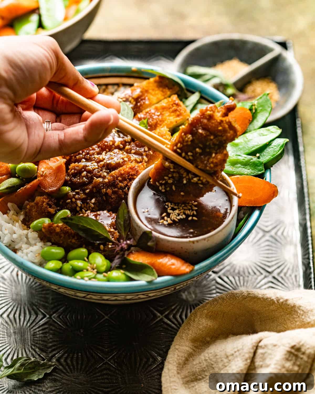 A hand holding chopsticks dipping a piece of crispy chicken into teriyaki sauce.